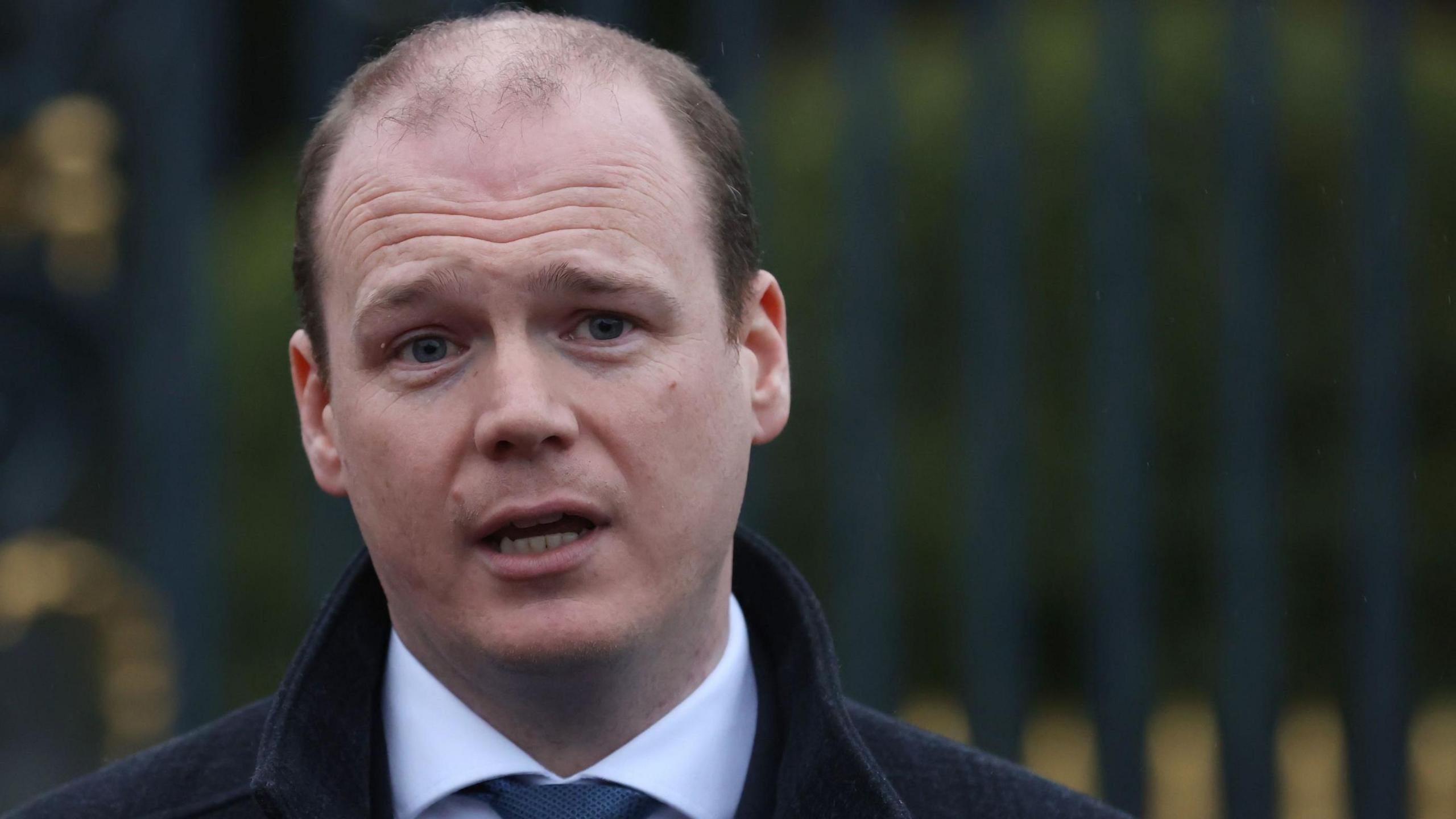 Gordon Lyons is standing with his mouth slightly open in front of some railings. He has thinning brown hair and is wearing a shirt and tie with a woolen coat.