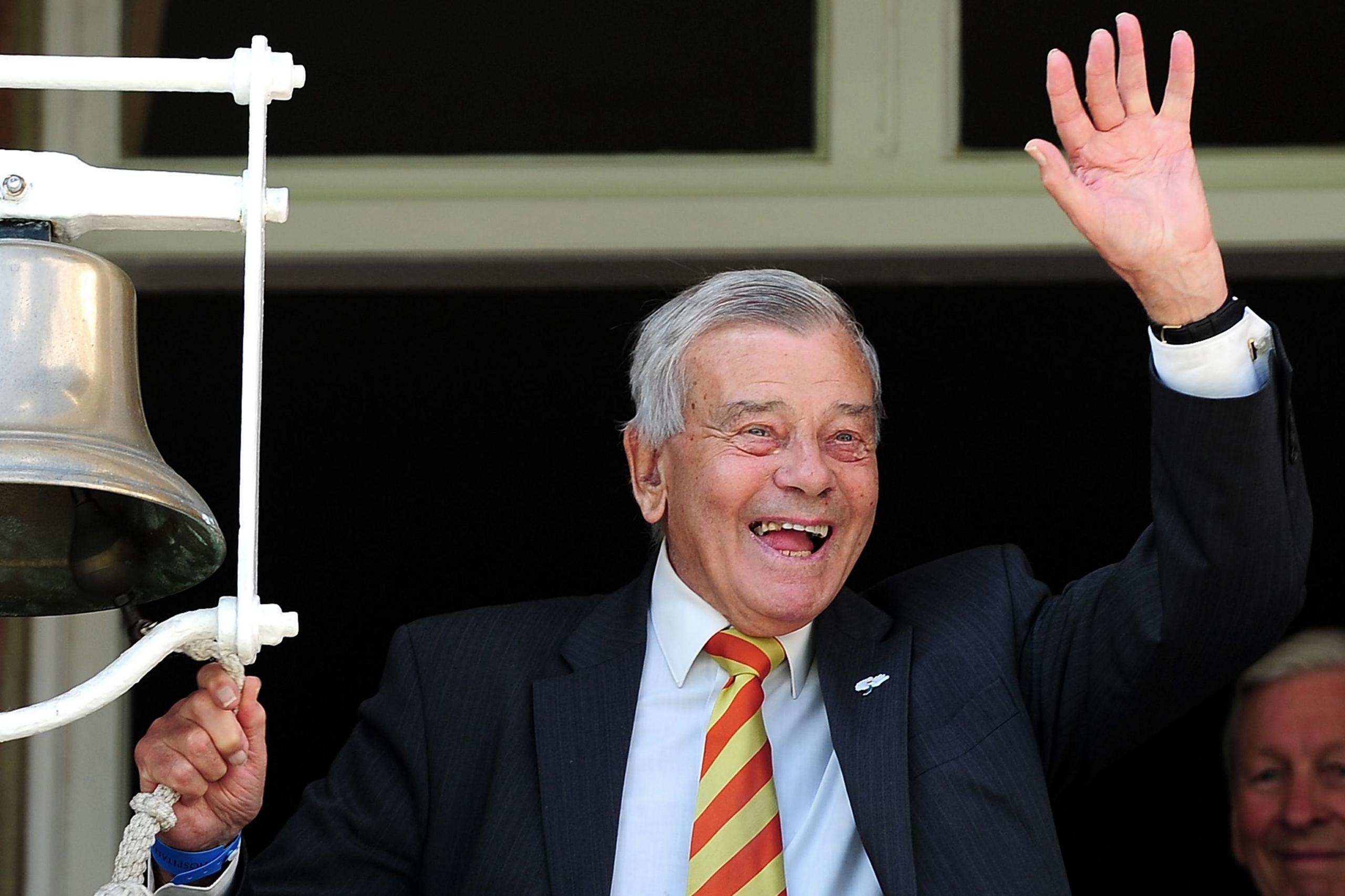 Dickie Bird, e veshur me një xhaketë të errët kostumi, një këmishë të bardhë dhe një kravatë me vija të kuqe dhe të verdhë, qëndron pranë një zile të madhe argjendi të montuar në një kornizë të bardhë. Njëra dorë mban litarin e ziles, ndërsa dora tjetër është ngritur në një gjest përkuljeje. Sfondi tregon një pjesë të një strukture ndërtese me një kornizë dritareje.