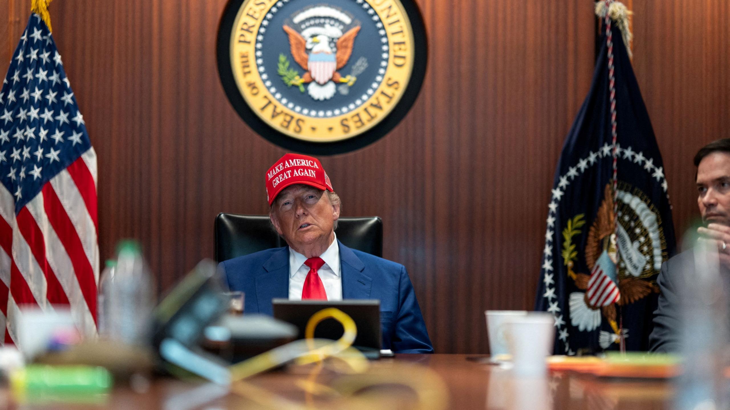 US President Donald Trump sits at a large wooden desk beneath a large presidential seal on the wall. He wears a "Make America Great Again" hat.