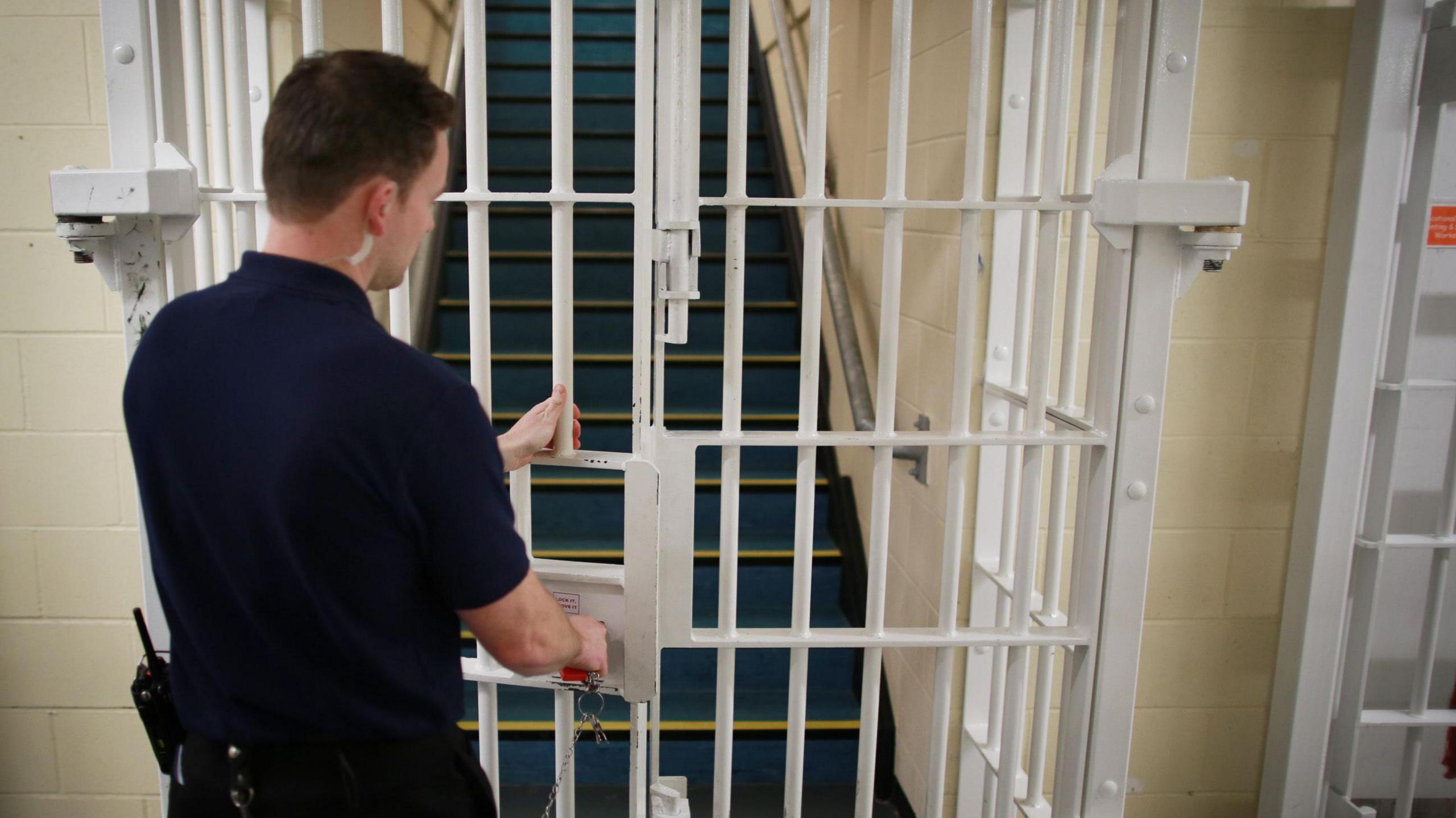 A police warden is closing a white gate inside a prison, with a set stairs behind it.