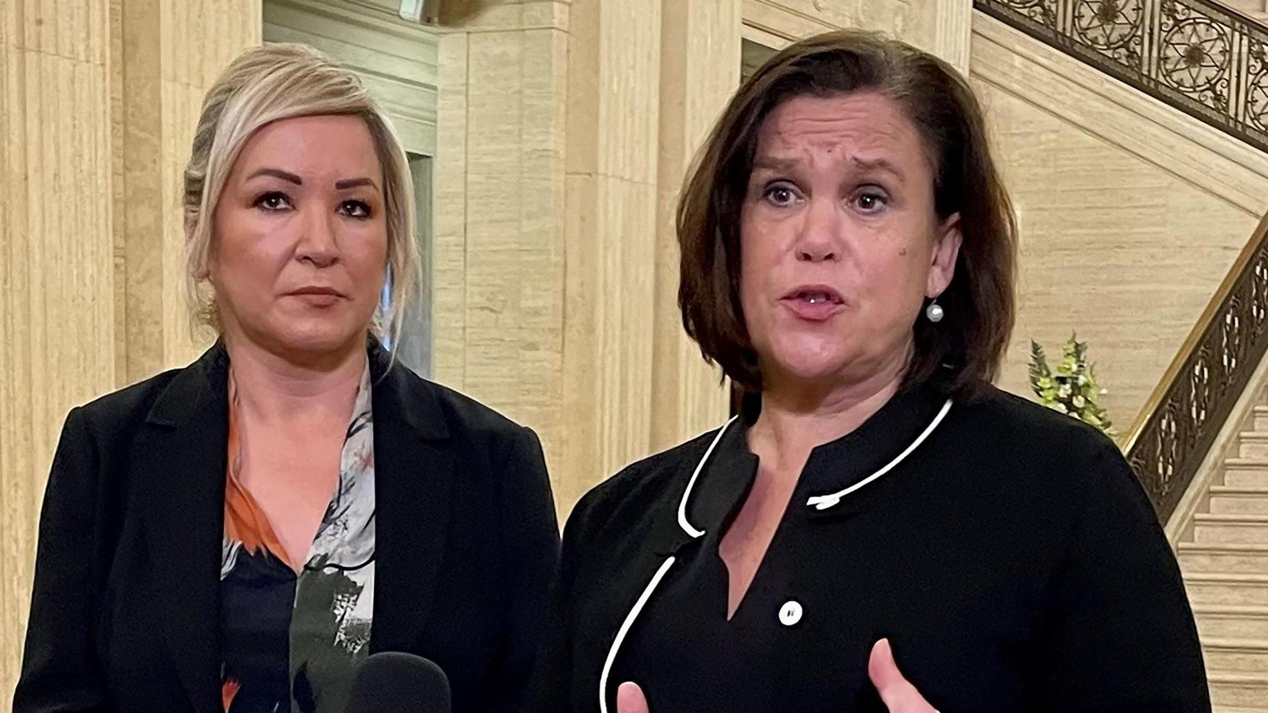 Michelle O'Neill has blond hair tied in a ponytail and is wearing a black suit jacket and patterned blouse. Mary Lou McDonald has short dark hair and is talking while wearing a black suit jacket