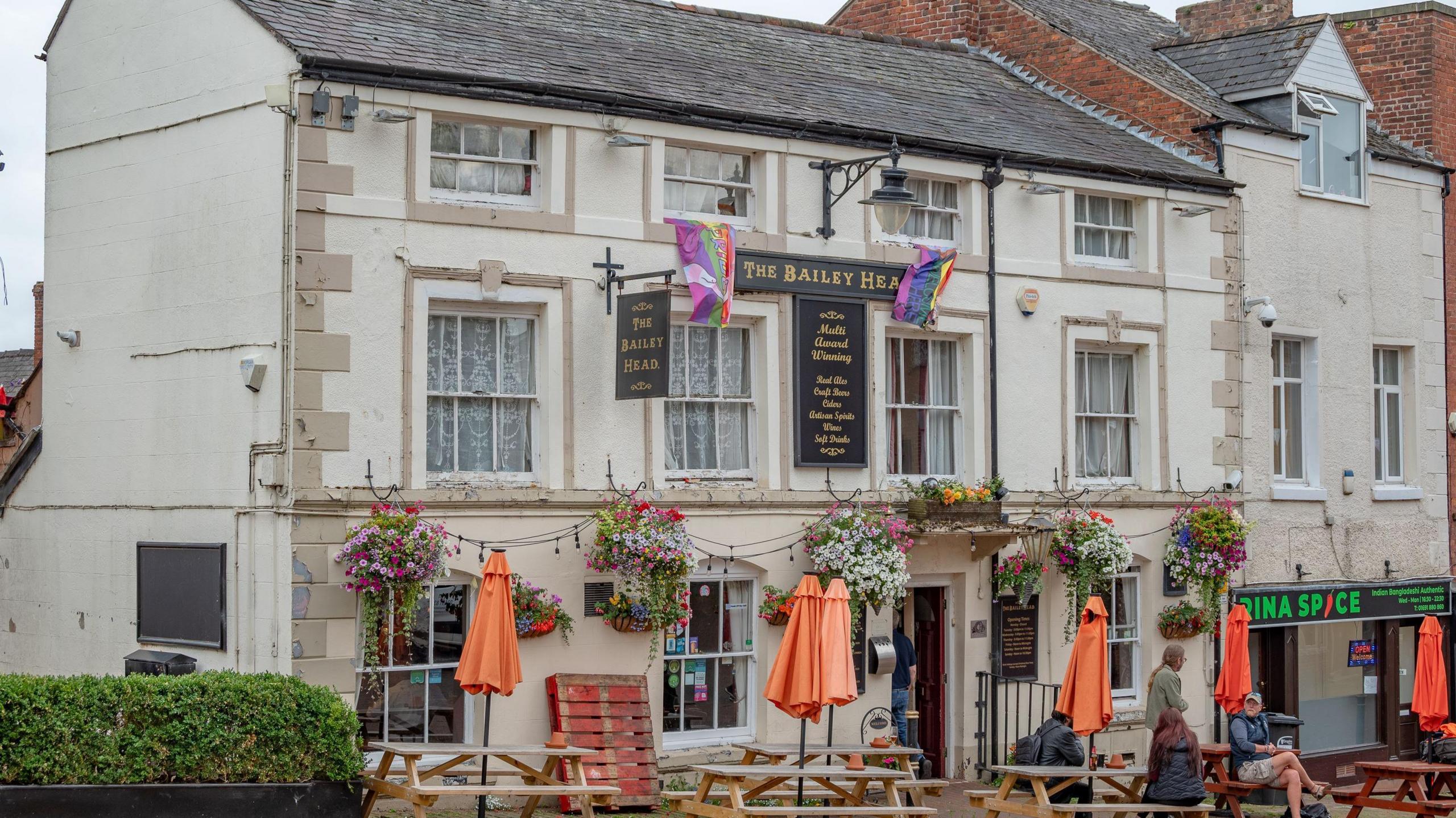 A three-storey white-coloured traditional pub with The Bailey Head on a sign above the entrance. Tables and benches are placed outside with people sitting on some of them.