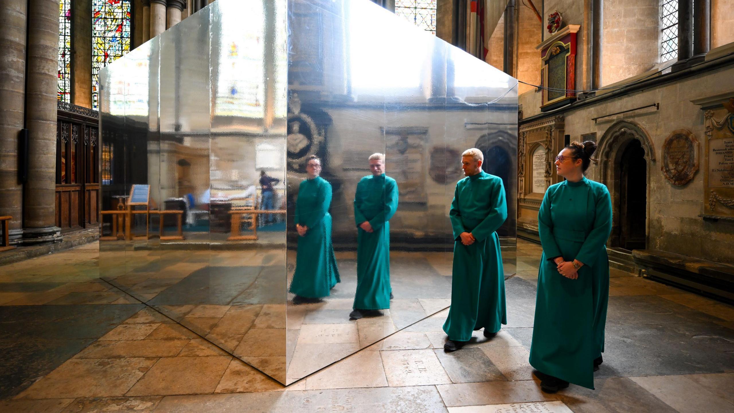 Two church clergy in green robes stand next to a mirror-coated box inside Salisbury Cathedral, in which their reflections are visible. It is part of the Living Water summer exhibition in Salisbury Cathedral