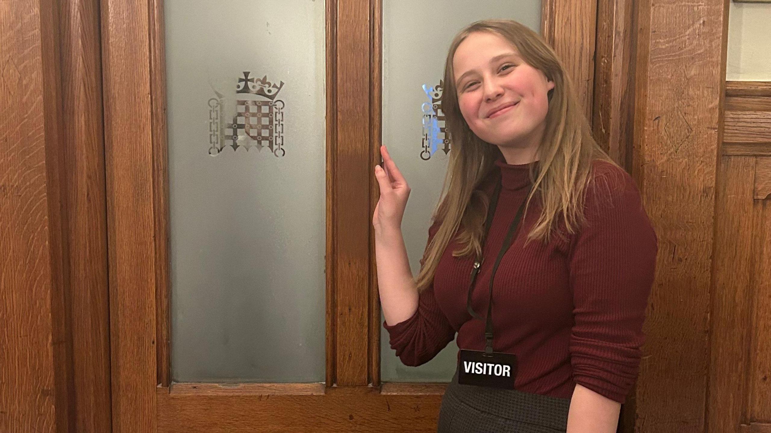 A young woman smiles at the camera. She is standing in front of a door with the Parliament logo on the doors. She is wearing a lanyard with an ID card reading "visitor".