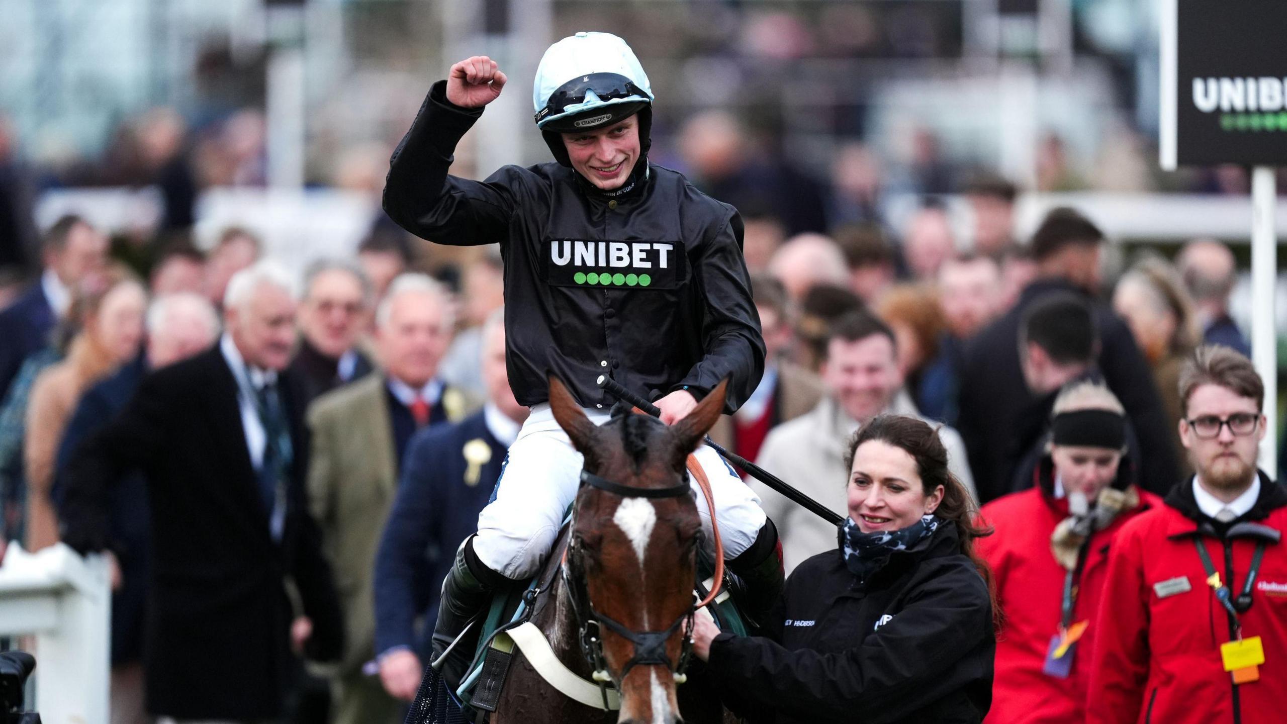 James Bowen holds a fist in the air as he is guided while seated on horse Holloway Queen with a crowd behind him at Cheltenham Festival