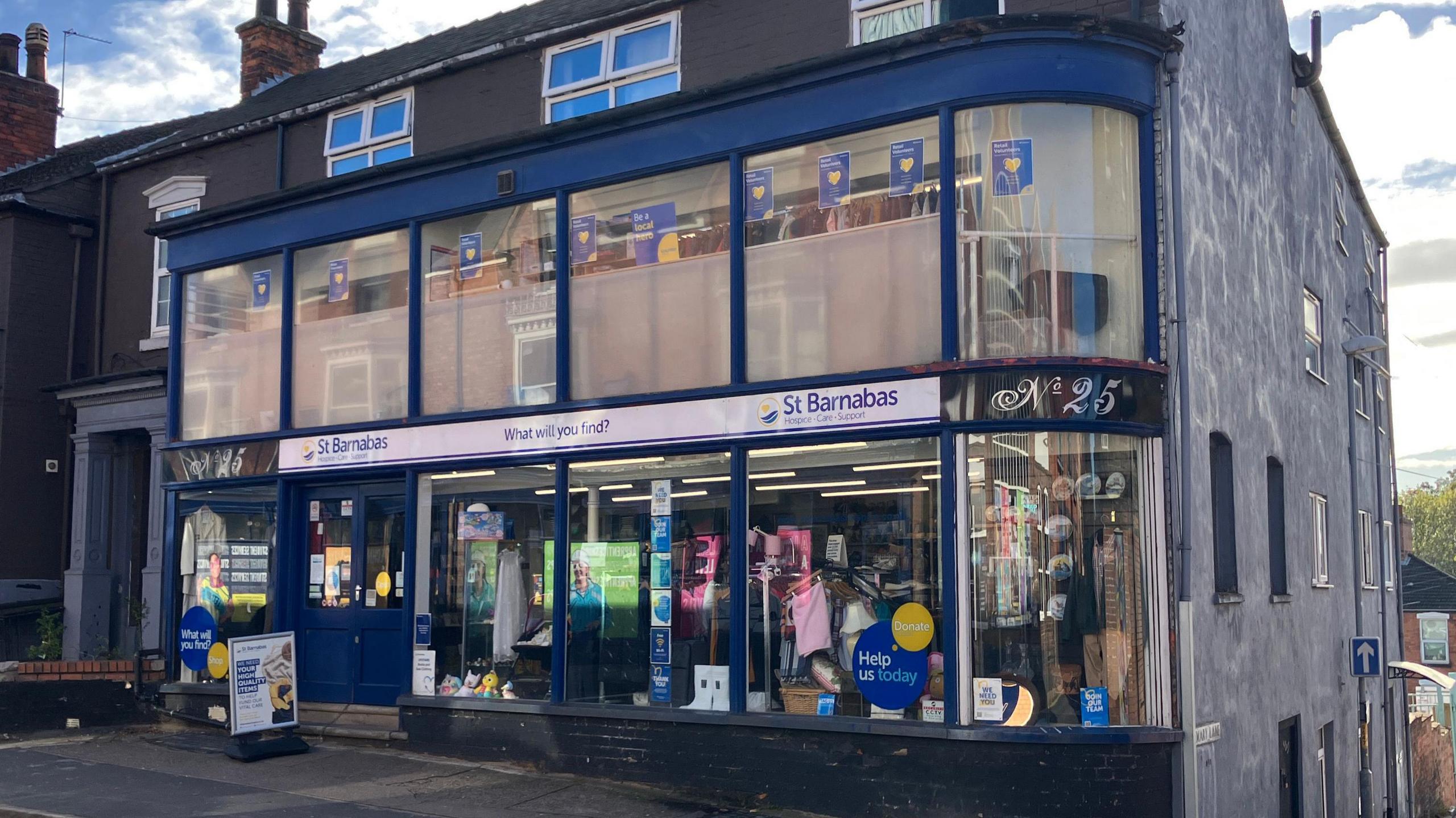 The outside of a blue charity shop called 'St Barnabas'. The shopfront has a navy frame and large glass windows both up and down. Inside there are mannequins clothes and shelves with bric-a-brac in the bottom windows. There is a white sign on the pavement outside. To the right, there is a black sign reading No 25. To the side of the shop are houses, and a large wall painted grey.