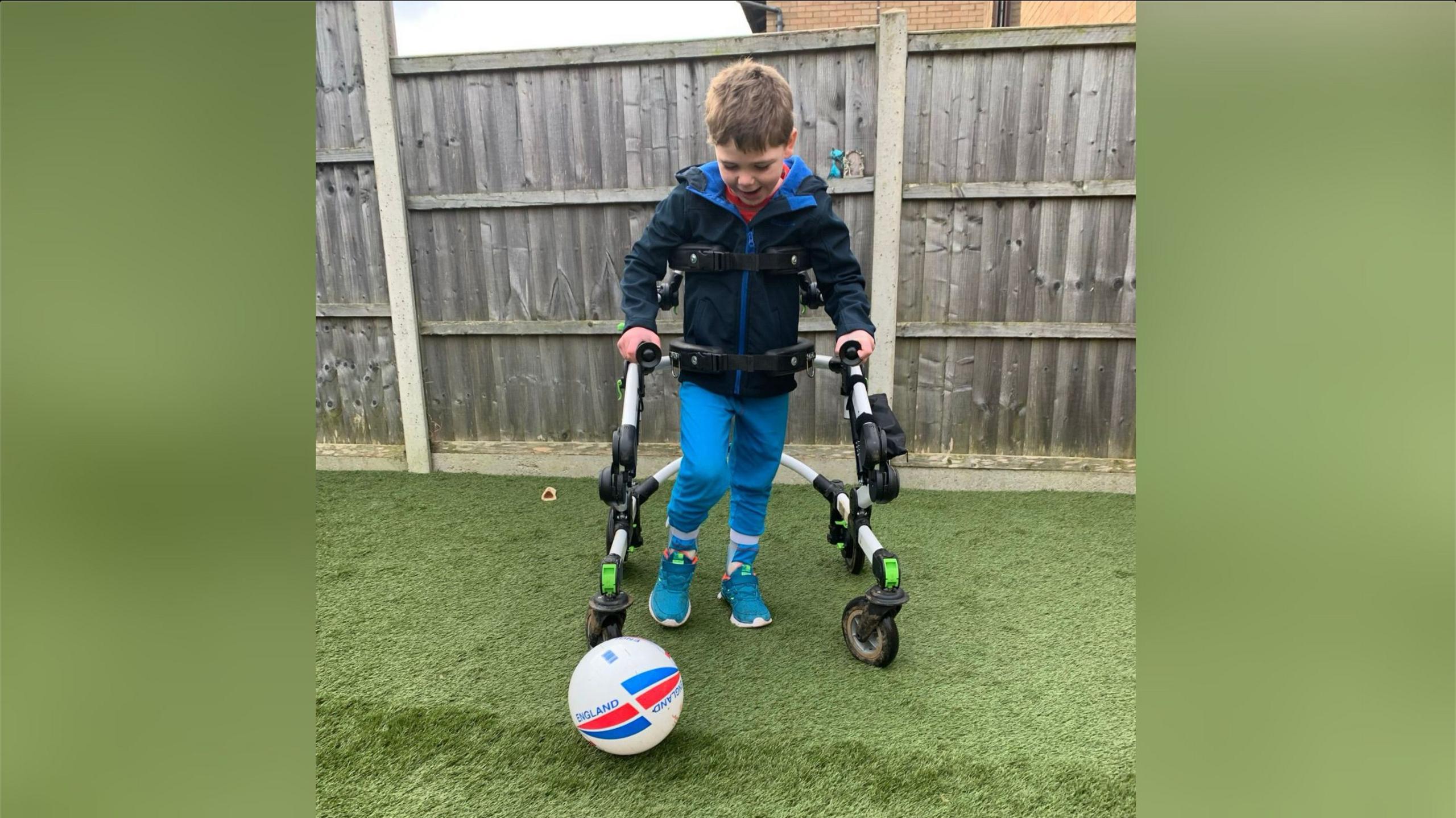 Isaac Tilley with short fair hair walking with a walker and kicking a football in a garden with a wooden fence behind.