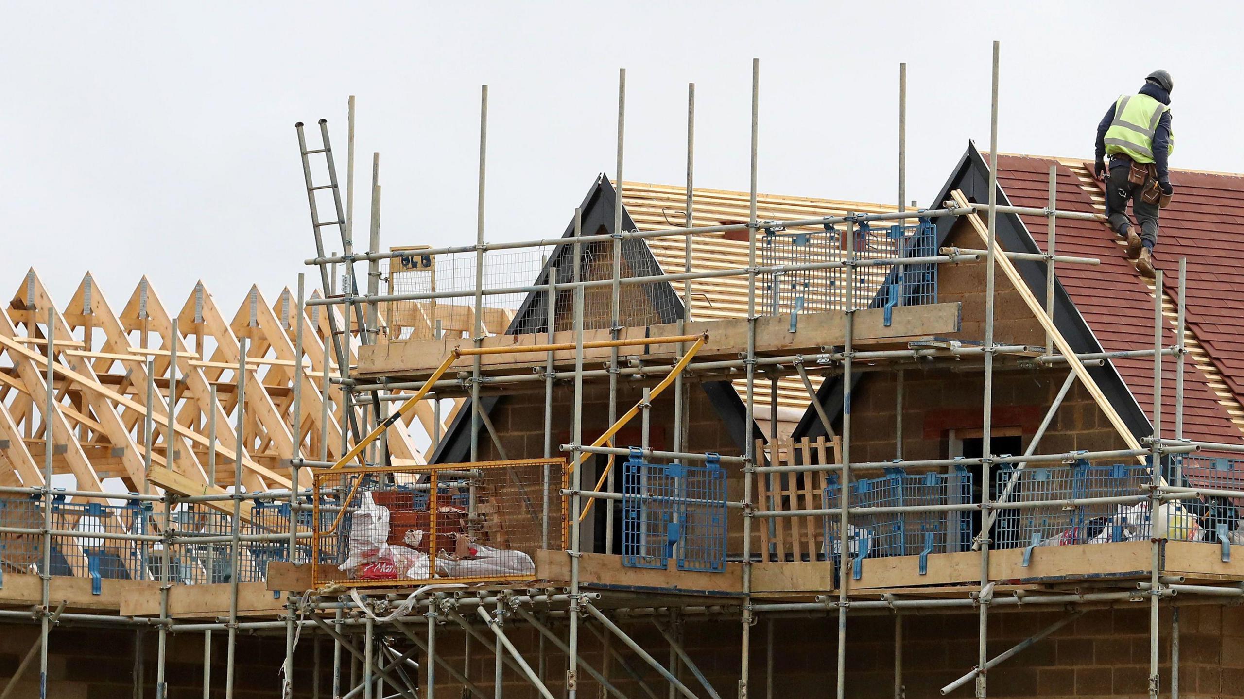A general view of new homes being built. The roofs are covered in scaffolding. A lone worker wearing a hard helmet and high vis jacket works on one of the roofs.