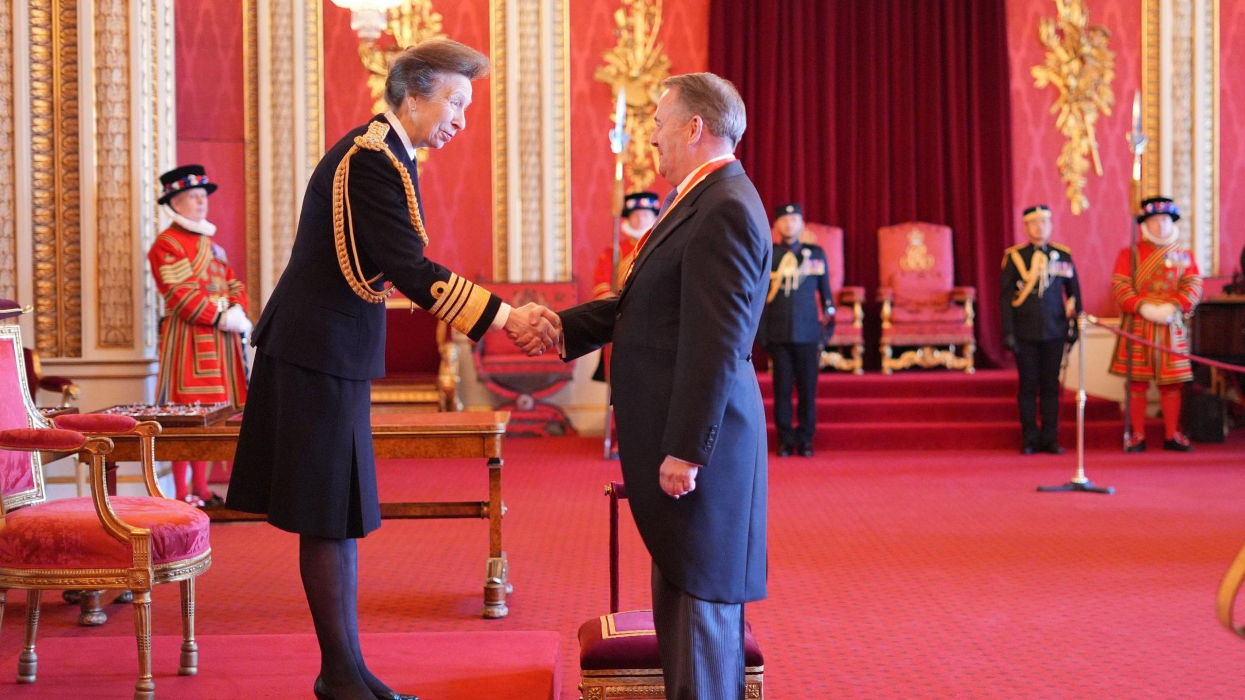 Anne, Princess Royal, is shaking Sir Liam Fox' hand at Buckingham Palace. The room is decorated in red and gold.