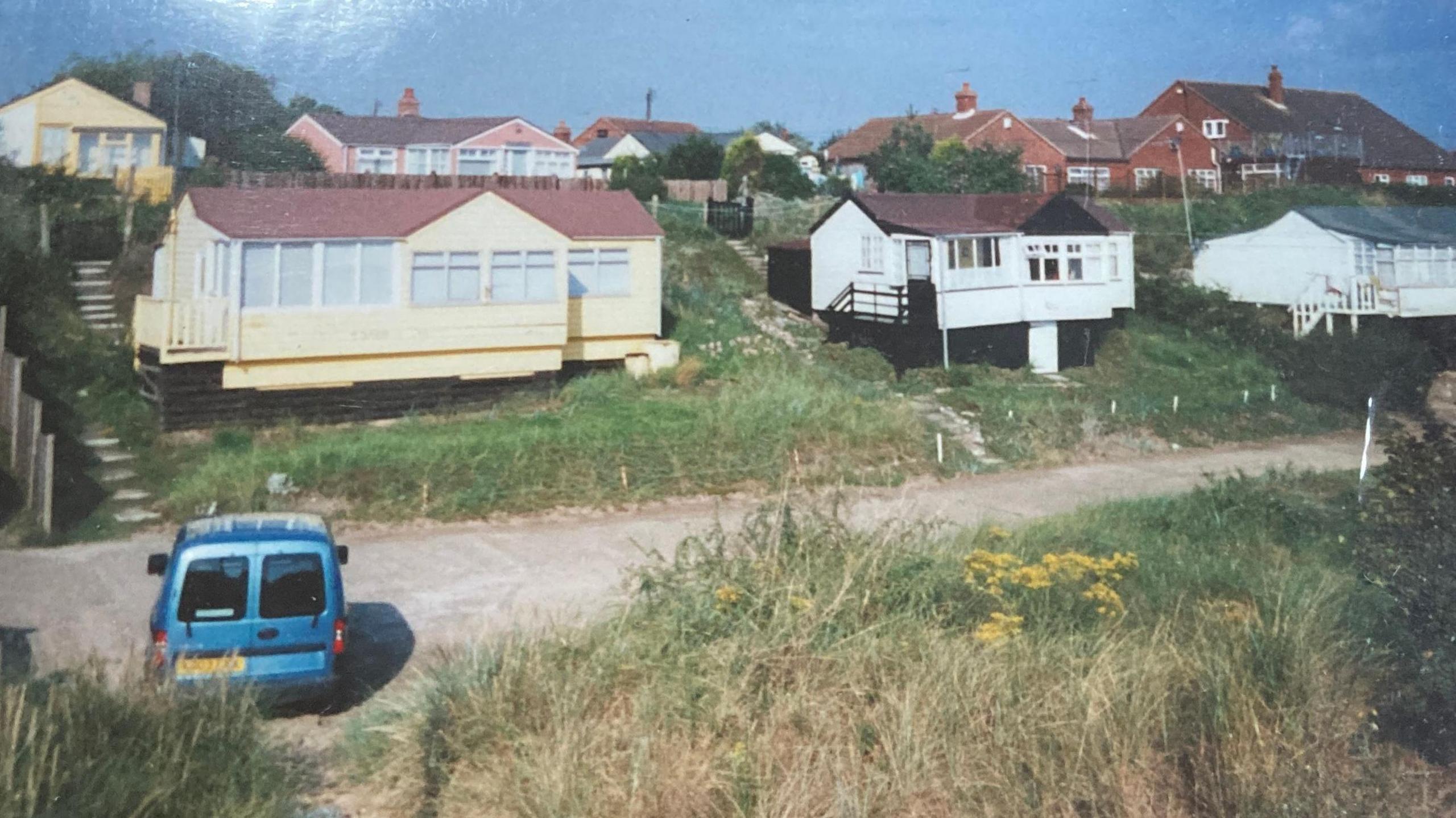 Three chalets with an access road in front of it and properties in the background. 