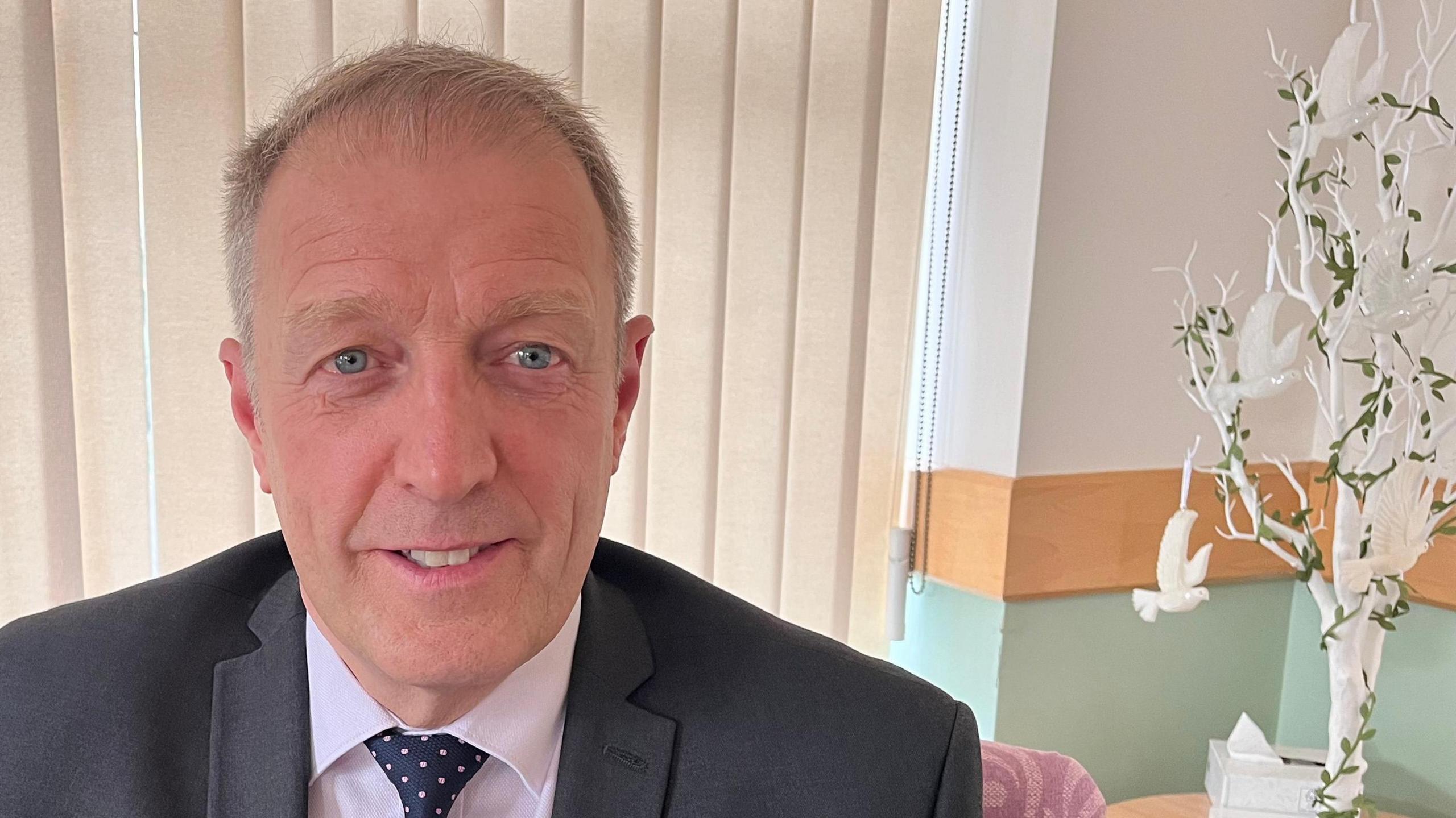 Chris Sadler sits in an office at Dove House Hospice. He has short grey hair and is wearing a grey suit, white shirt and blue and pink tie. To his left is a sculpture of a white and green tree white white doves.