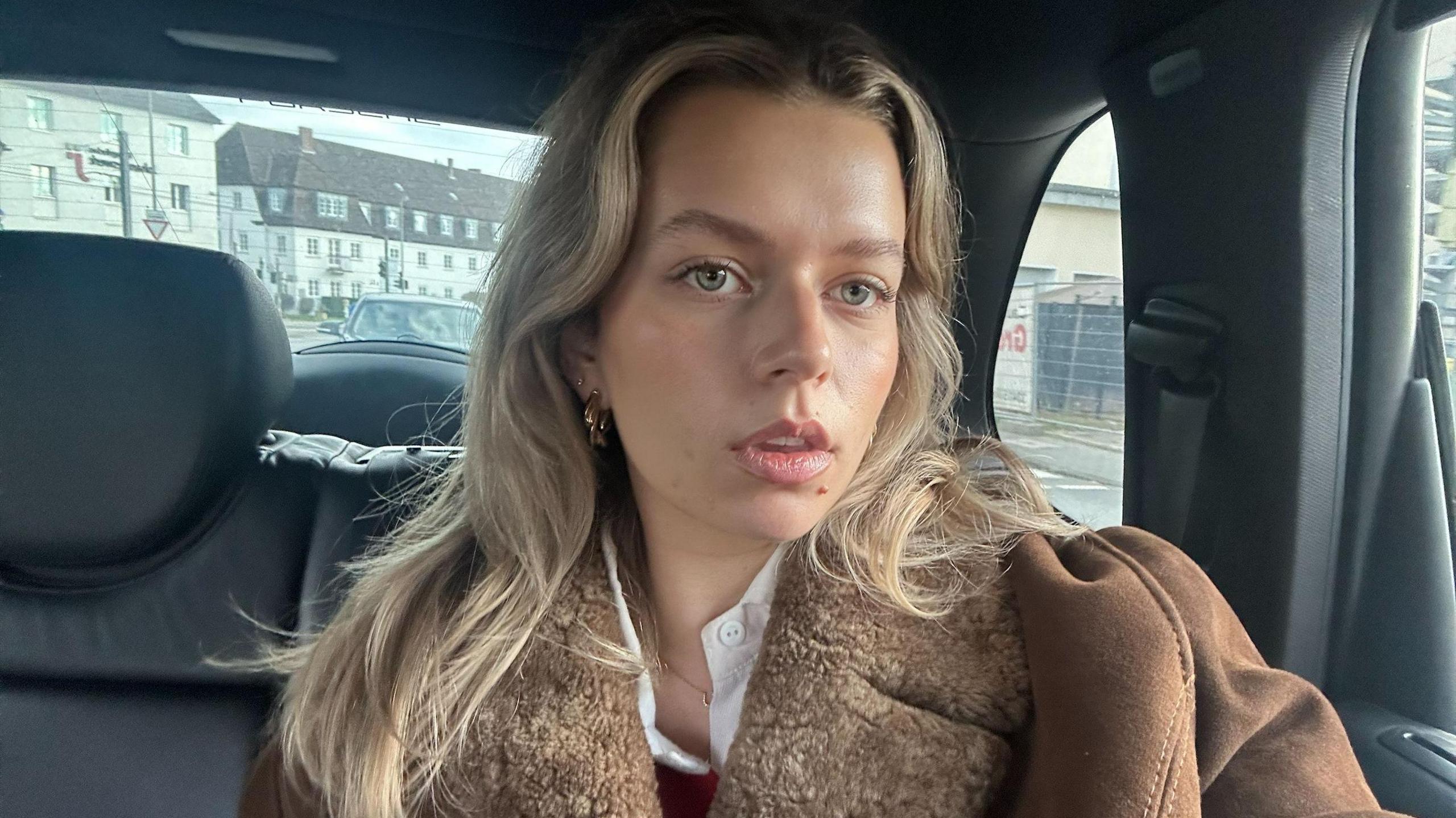 Sumi Mrkulic with long blonde hair in the centre frame of a car, she is wearing a brown coat and gold earrings