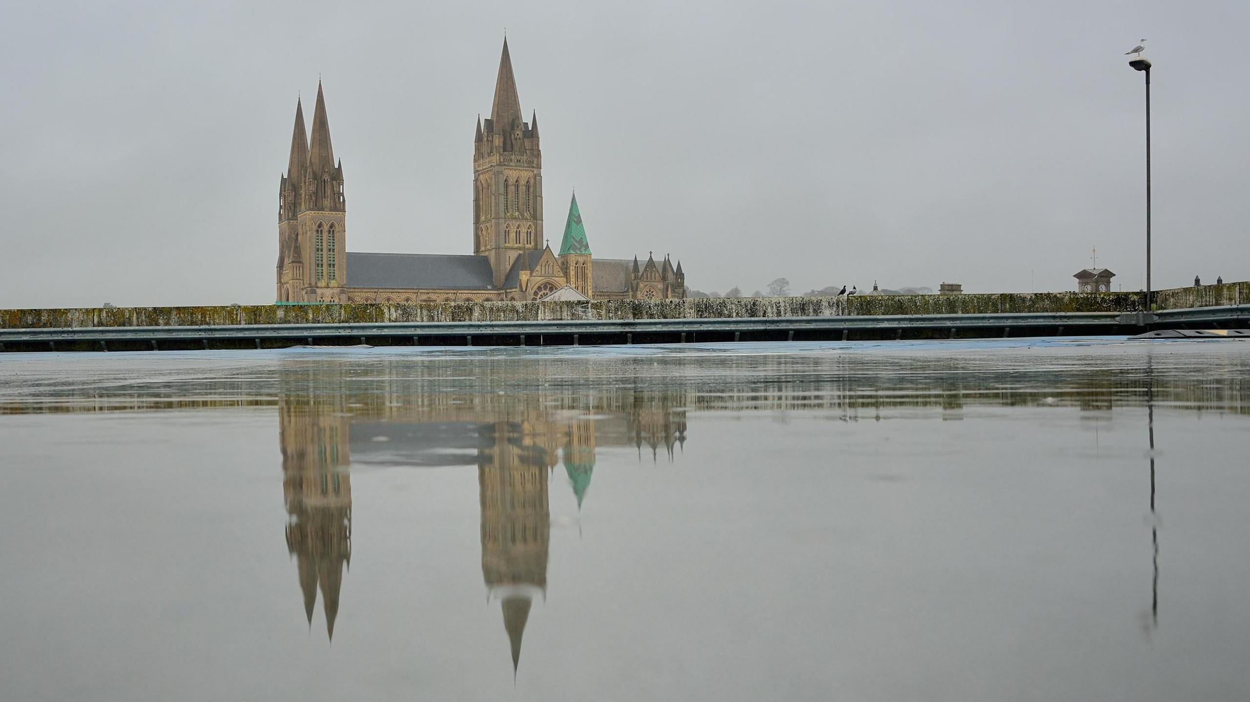 A gloomy day in Truro. Truro Cathedral is in the distance. Its reflection can be seen in the water from rain which has settled on the surface of a car park.
