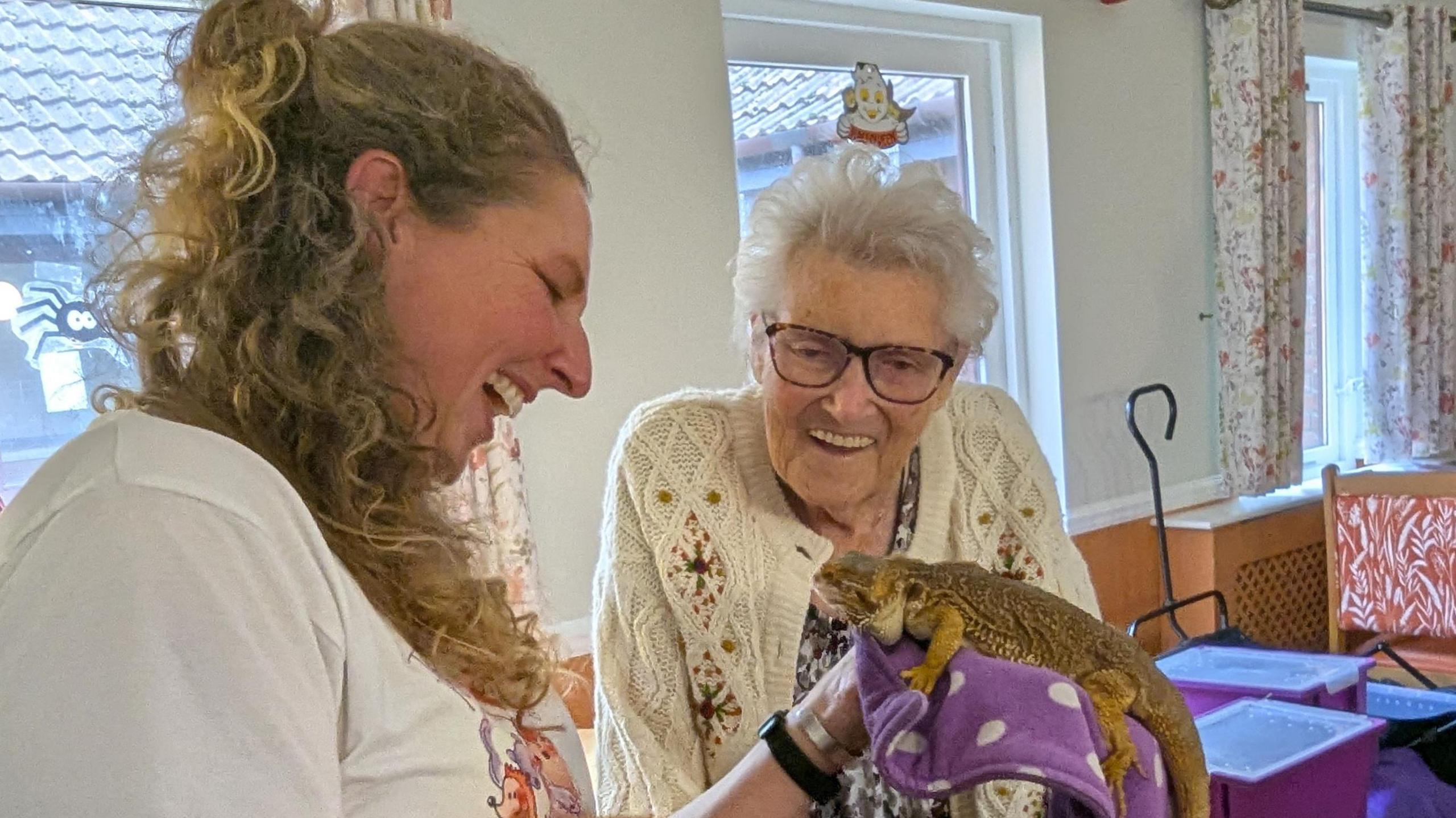 Two women are interacting with a bearded dragon lizard. The woman on the left holds the lizard on a purple cloth, while the other lady on the right looks down at it, smiling widely. The lady holding the lizard wears a white t-shirt and has blonde curly hair. The elderly lady on the right has short, white hair, wears large glasses and has on a white knitted cardigan with diamond detailing.