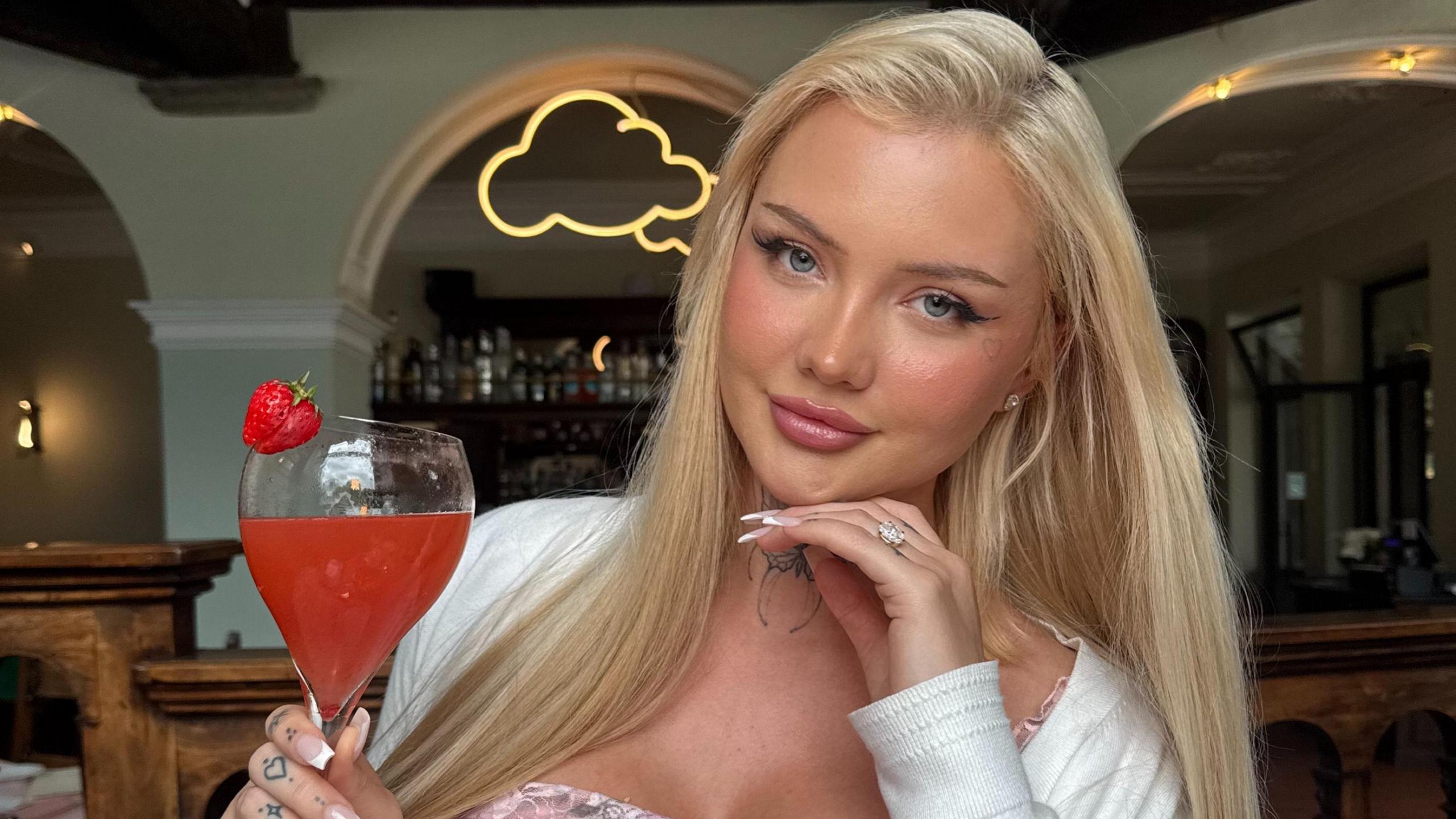 A woman with long blonde hair is smiling and holding a cocktail. She is wearing a pink dress and white cardigan.