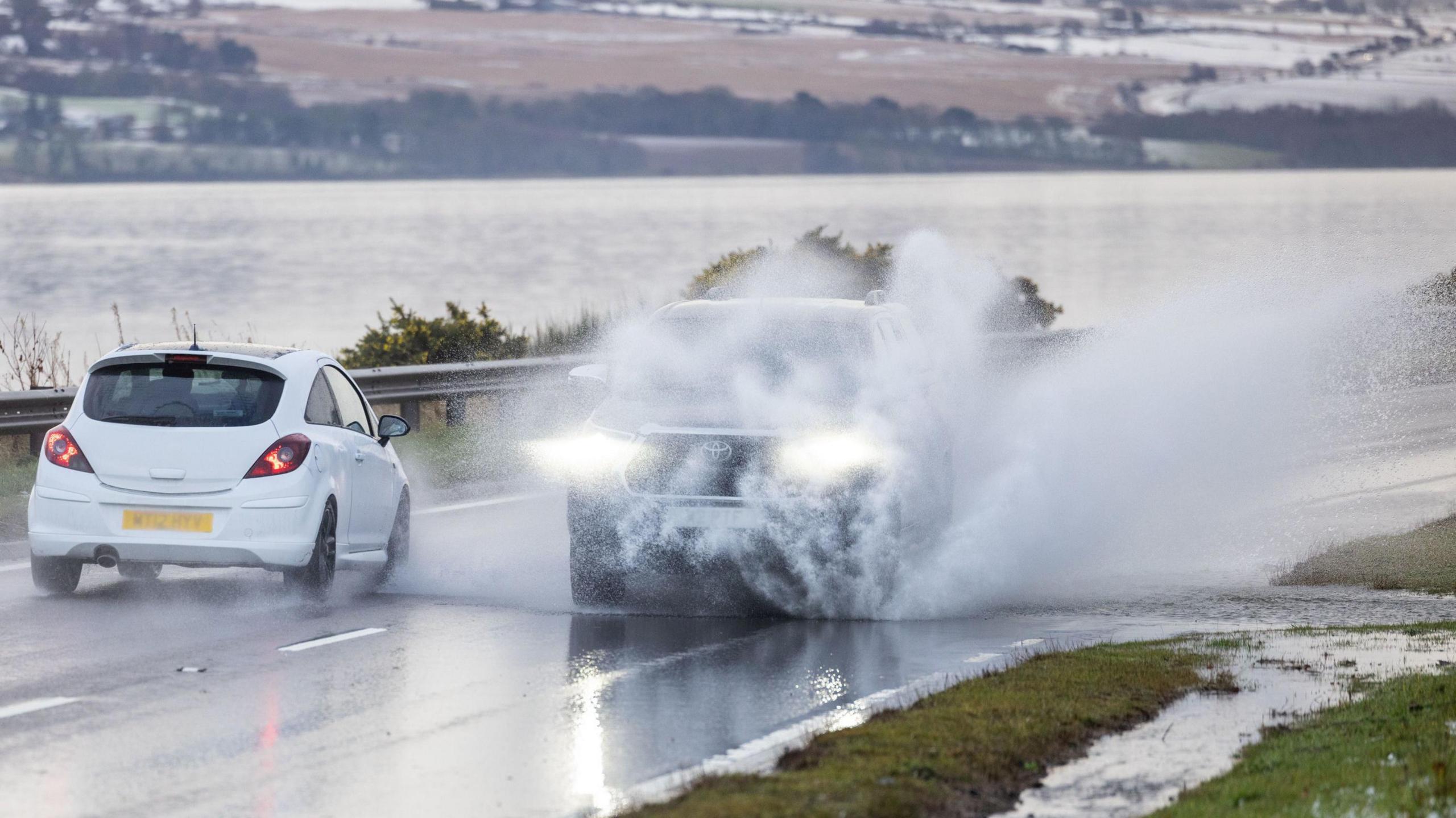 A Toyota 4x4 is covered in spray after driving through surface water on the A9 near Inverness. A small white car is driving past the vehicle, heading in the opposite direction. Snow can be seen in the fields in the background