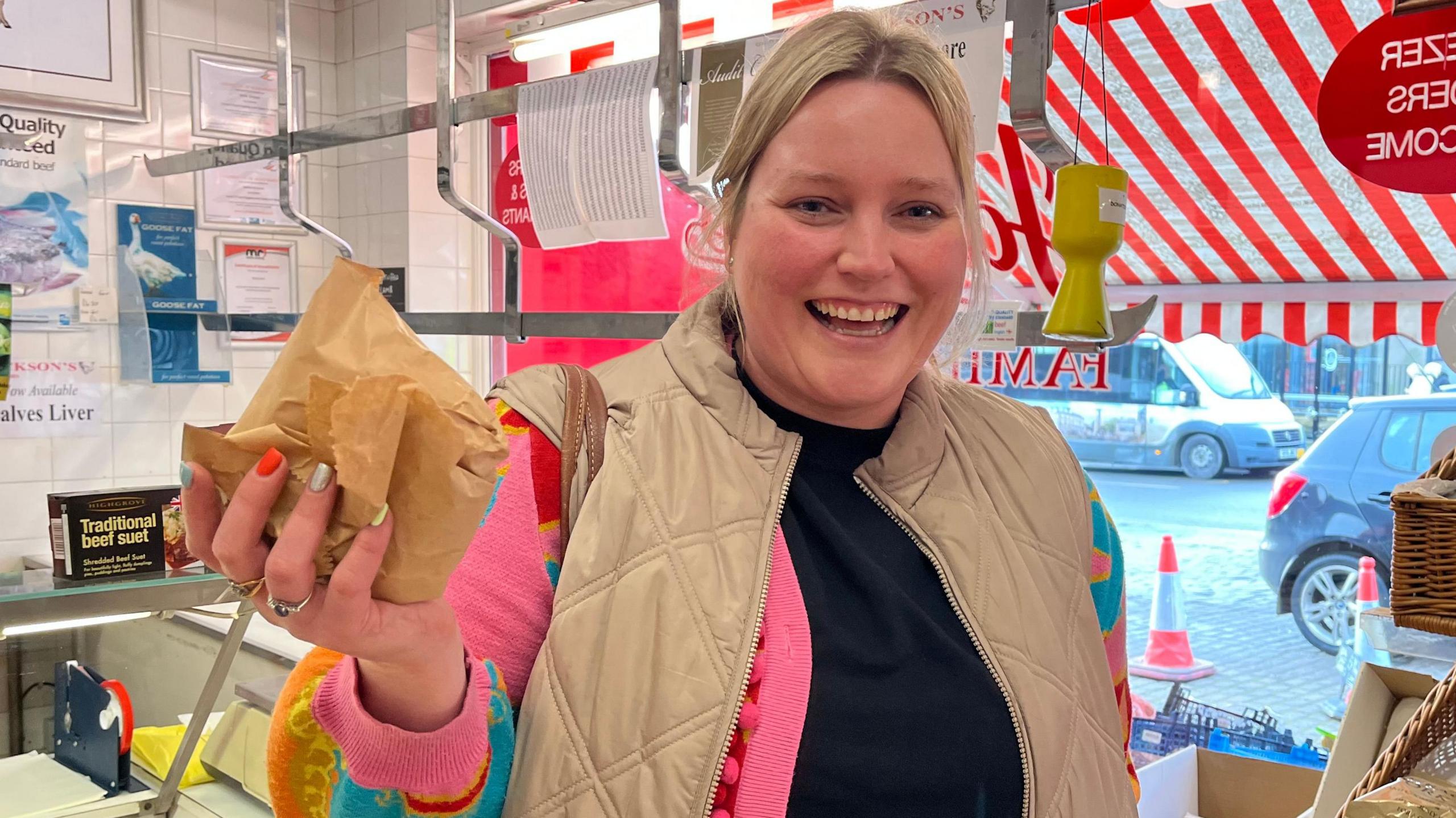 A woman with blonde hair, wearing a cream colour gilet and a pink cardigan is standing inside a butchers shop. She is holding up a paper bag.
Her nails are painted silver, orange and light blue.