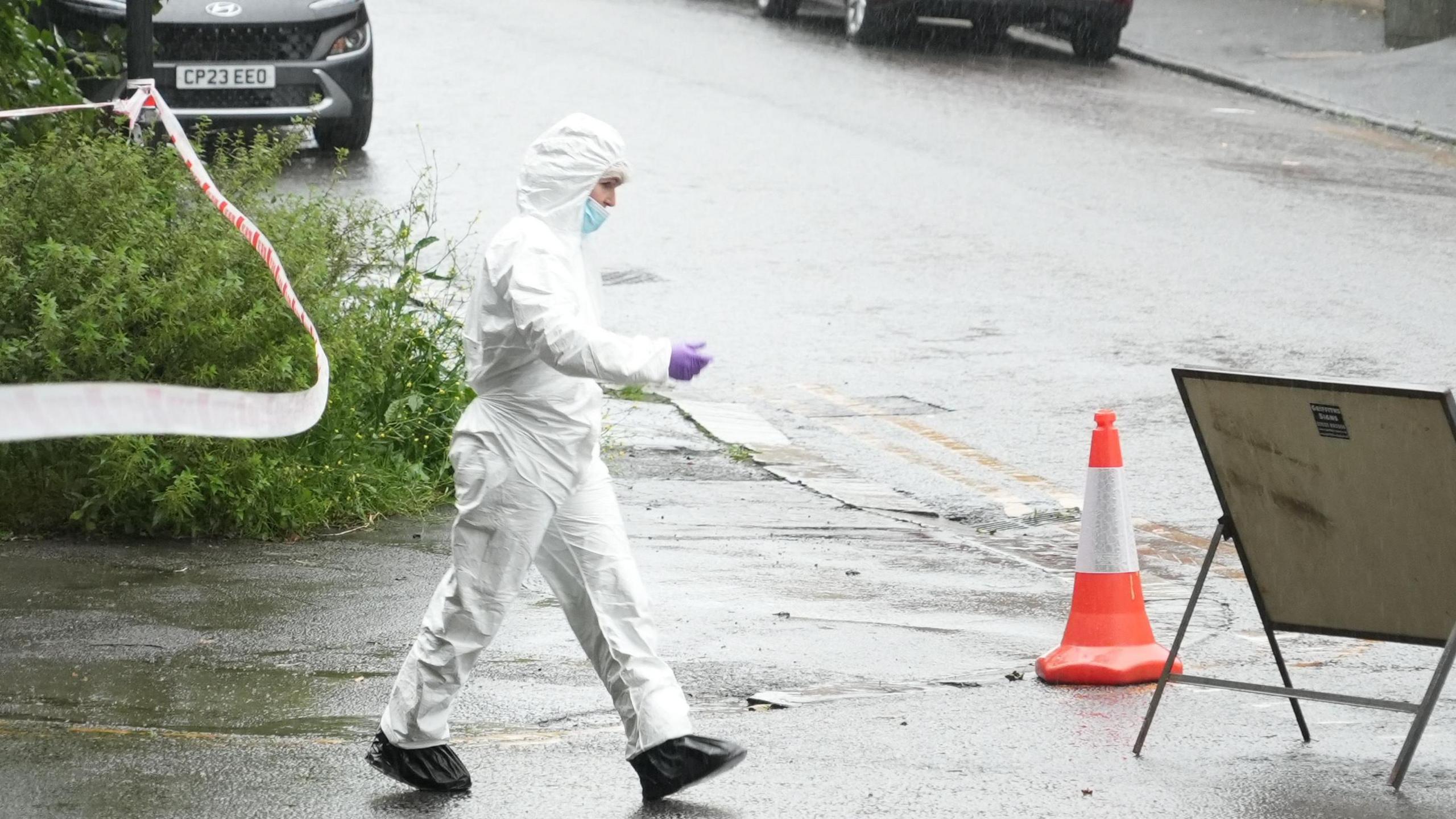 An officer in forensics kit, which is a white all-in-one suit and mask, on a rainy street with cordon behind them