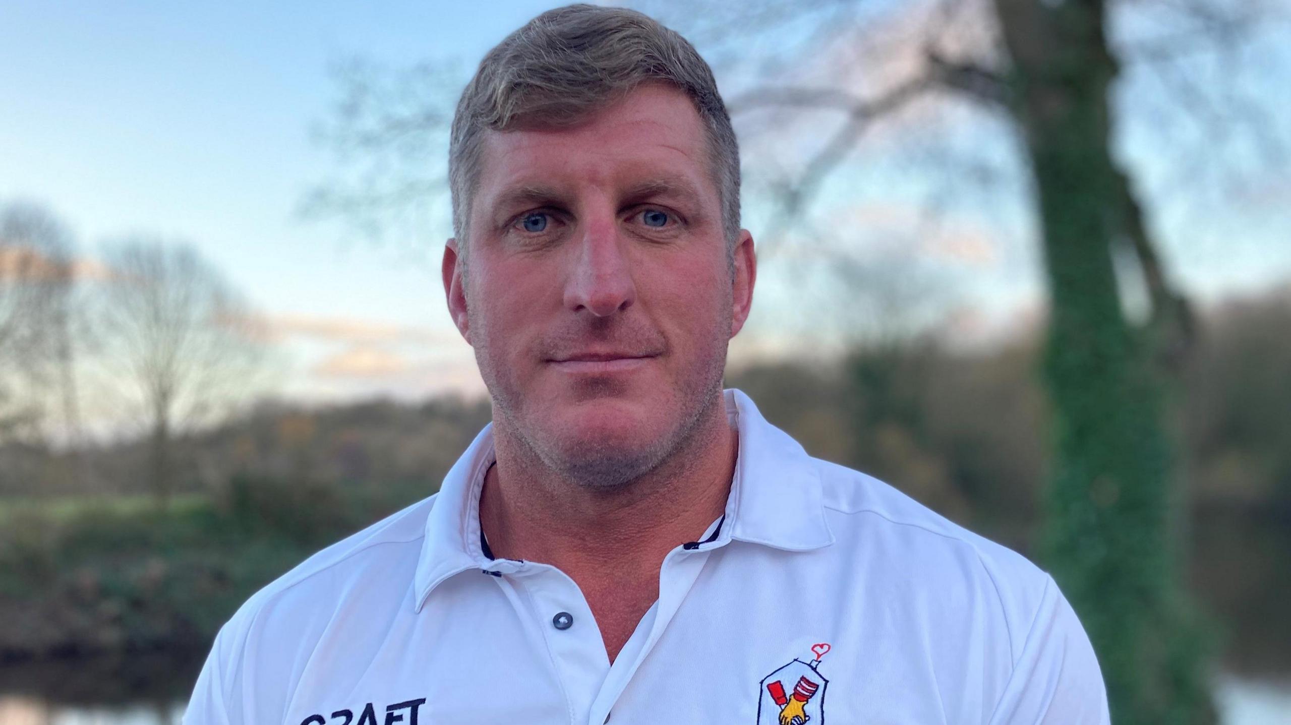 Dean Fitzmaurice has short sandy-coloured hair and is wearing a white polo shirt. Trees and water can be seen behind him.