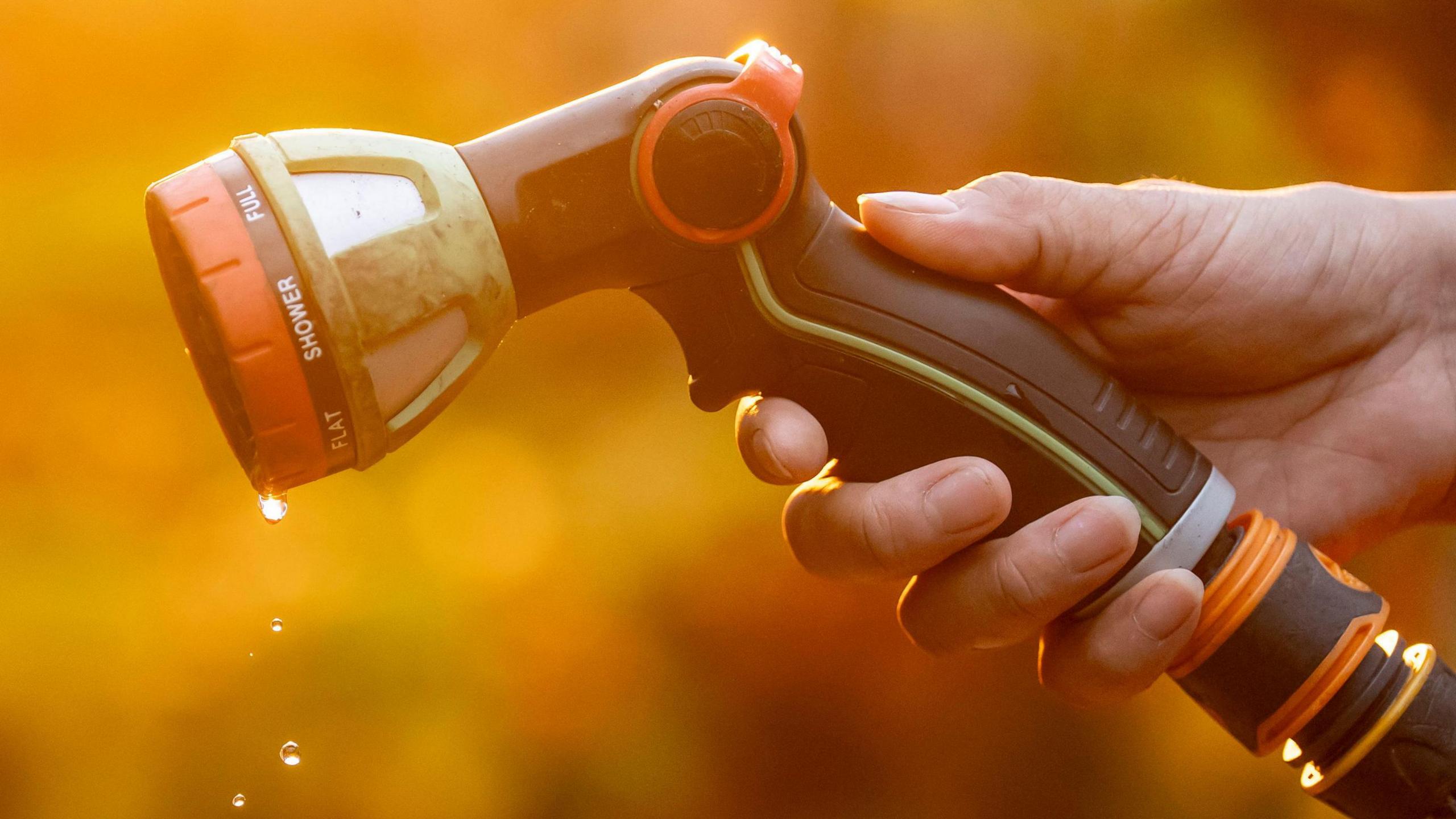 An orange tinted image of a hand holding a garden hose with water droplets dripping from it. 