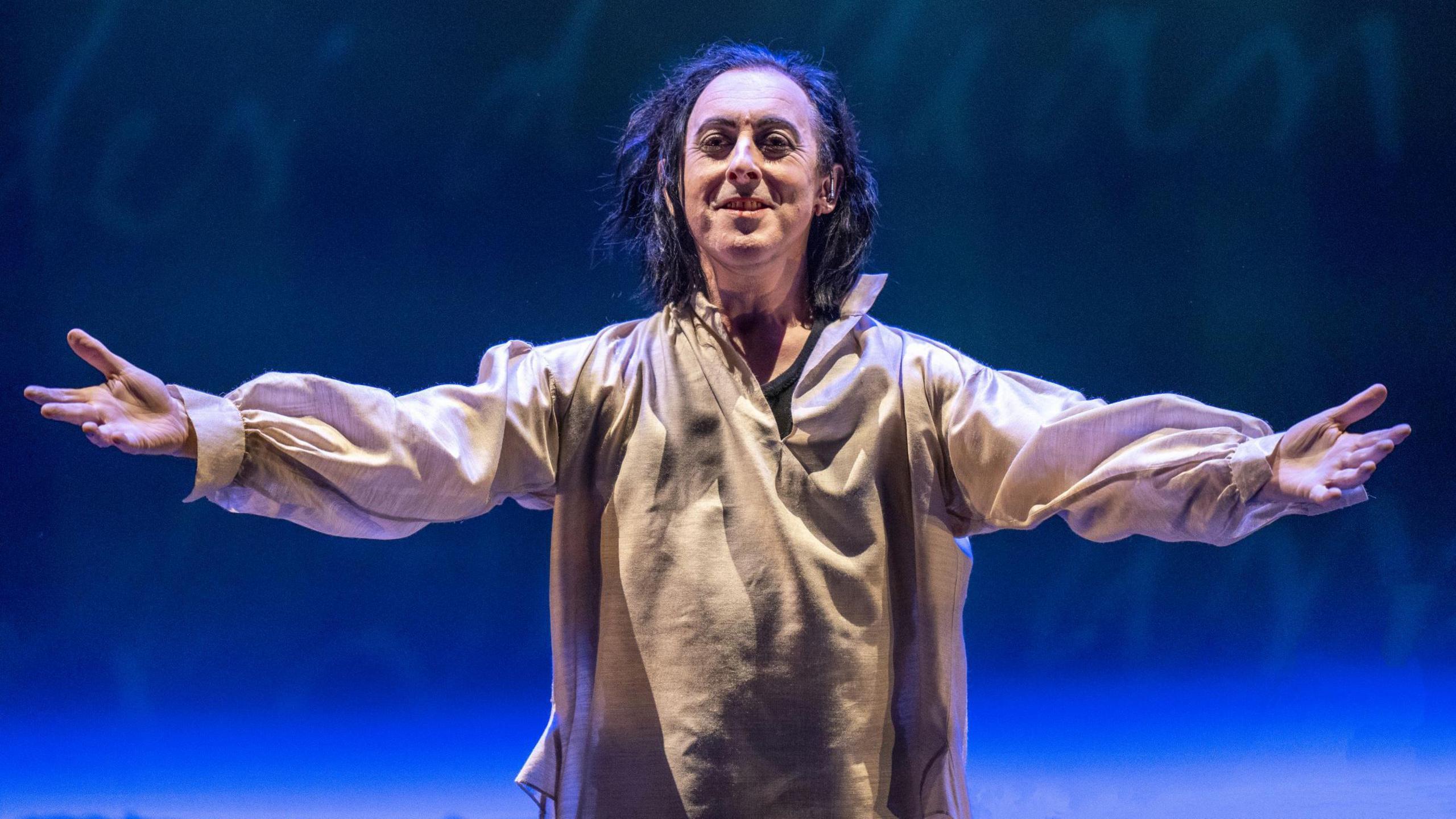 Alan Cumming on stage in Burn in 2022 - he has his arms out in a gesture that suggests he's welcoming people, while dressed in a somewhat tatty looking white outfit. 