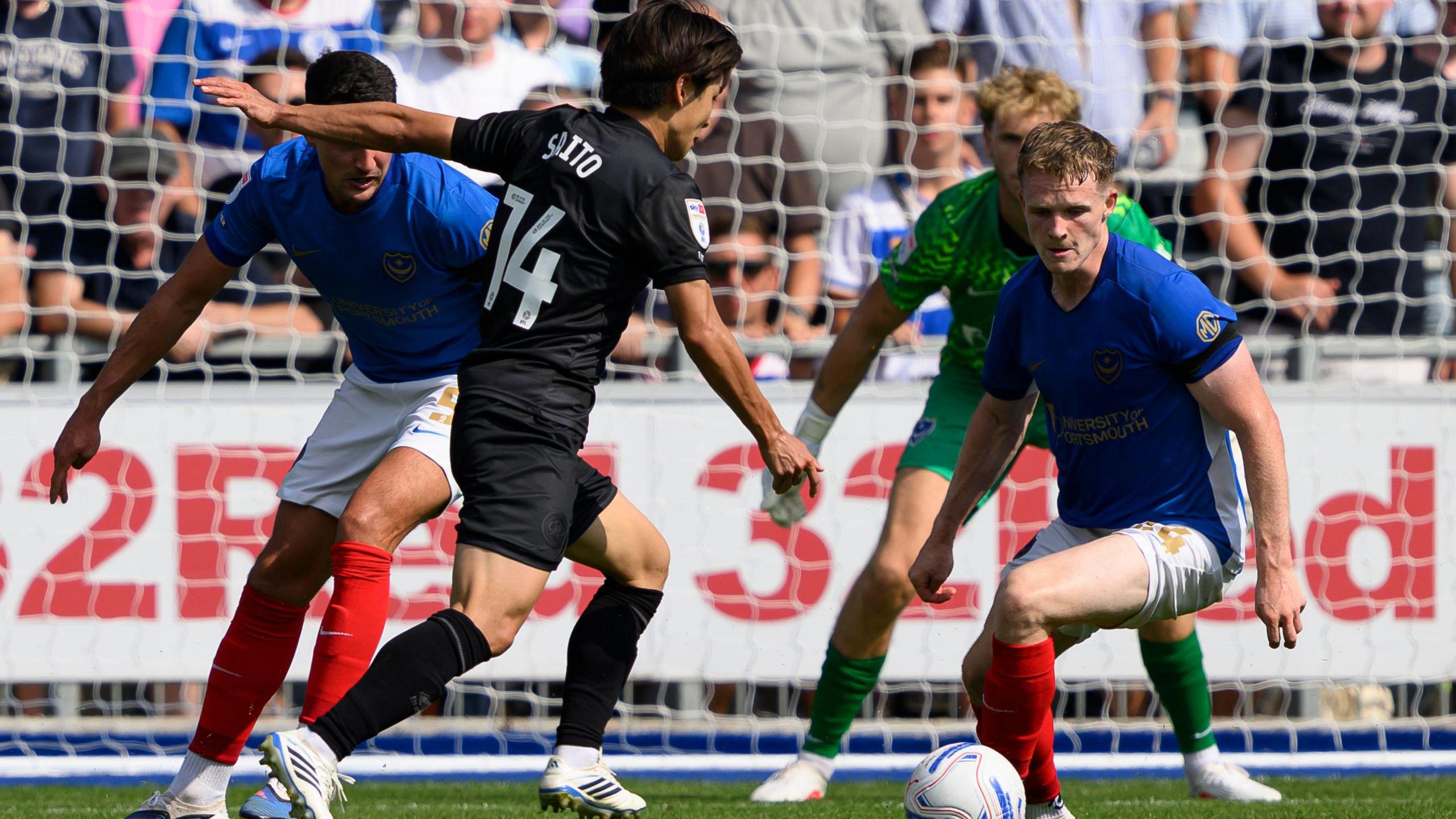 Koki Saito, wearing QPR's black away kit, moments before shooting to score against Portsmouth. The Portsmouth defenders, wearing their blue, white and red home kit can be seen trying to block his effort.