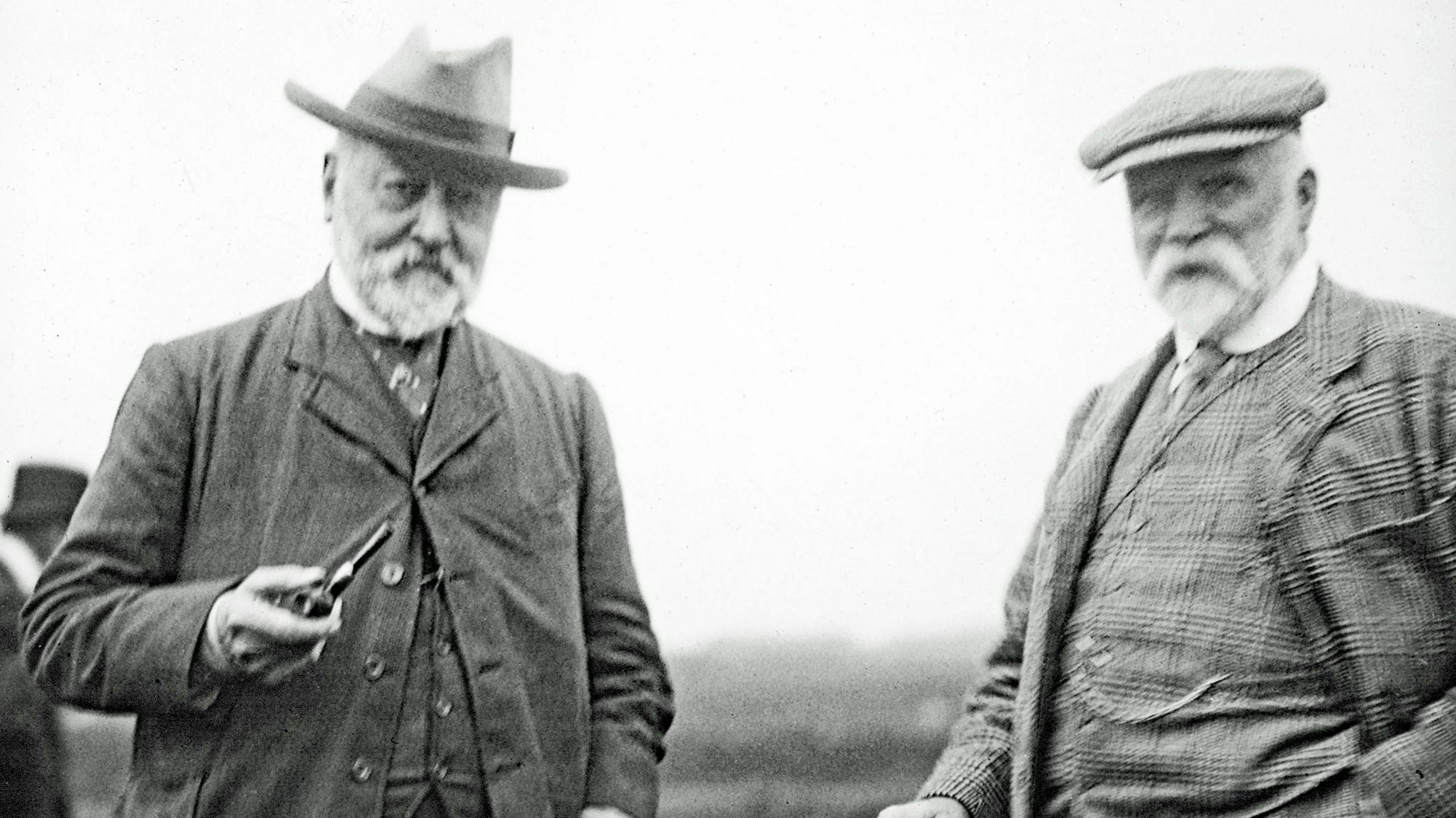 A black and white image showing the two men standing next to each. Both are older men with white beards. Alexander Ross is wearing a brimmed hat and a tweed jacket over a waistcoat. He is holding a pipe for smoking in one hand. The other man is also wearing tweed, and a flat cap.