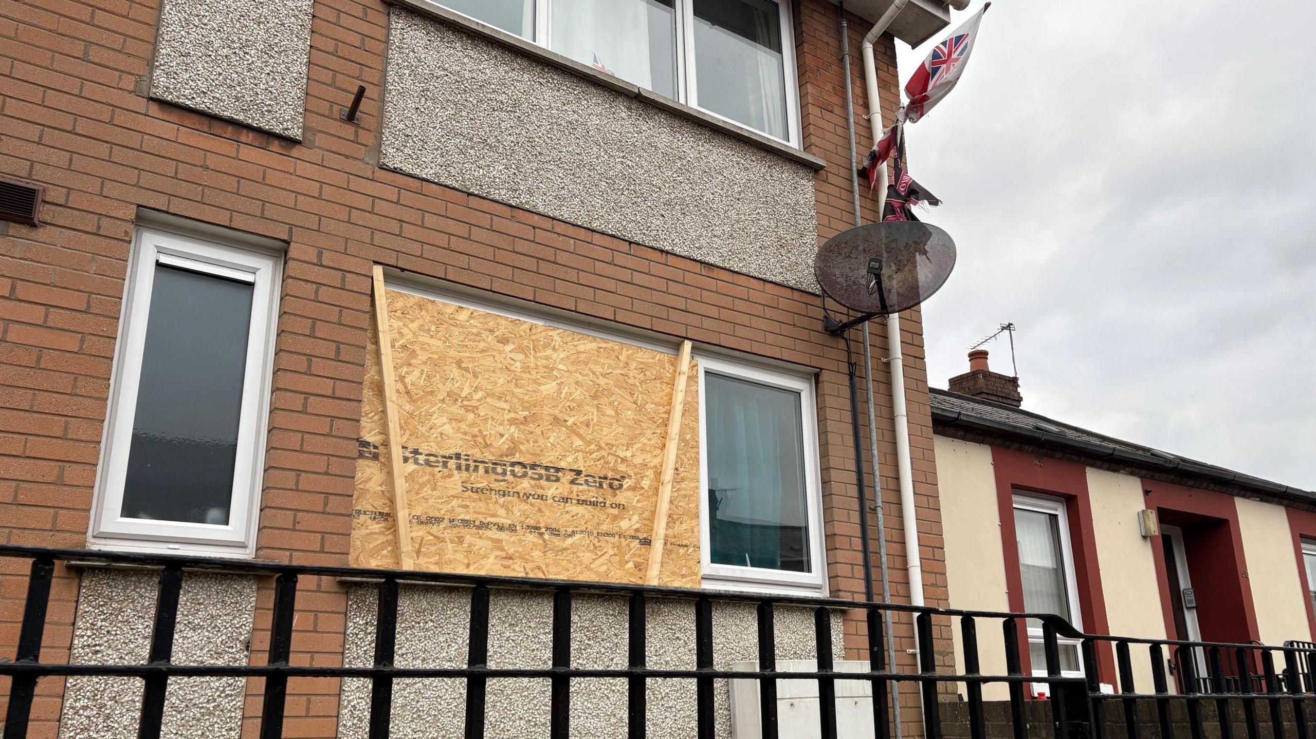 The ground floor flat in a beige brick complex in Templemore Street.  One of the front windows has been boarded up with plywood.  There is a satellite dish and a flagpole attached to the outside of the building. 