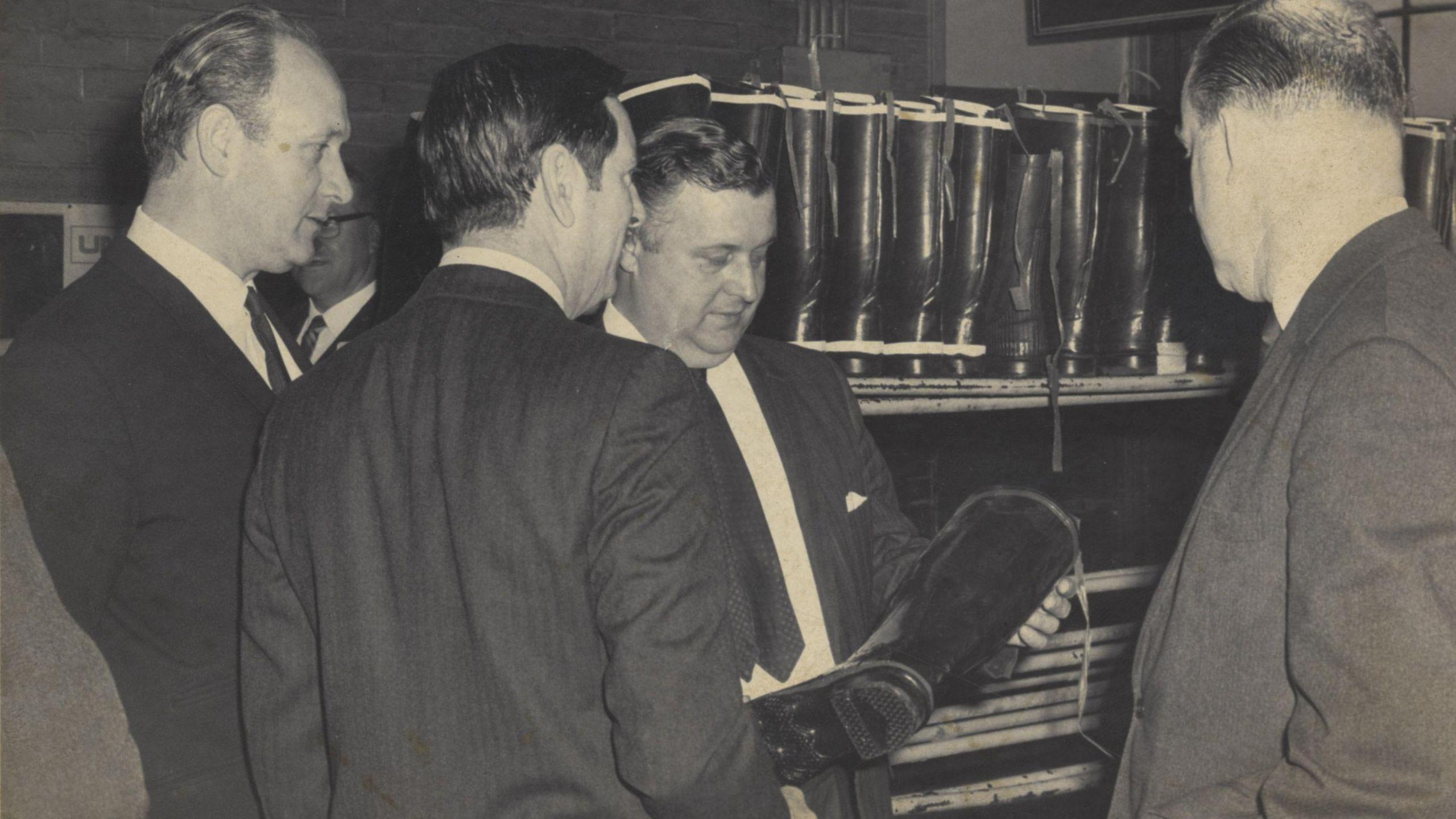 A group of men in suits and ties examine the quality of a wellington boot