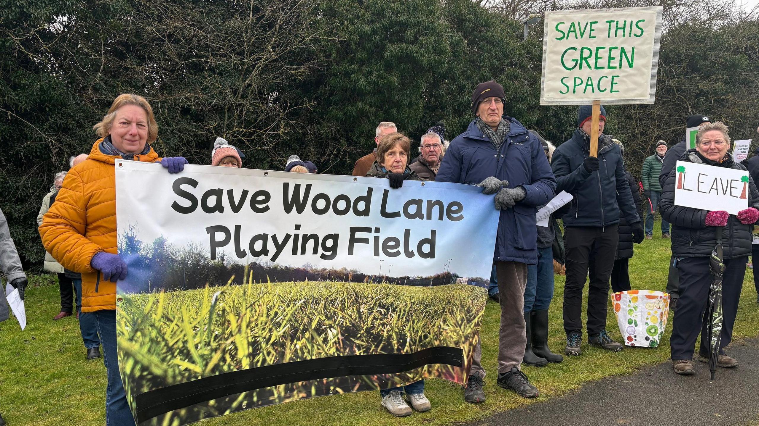 A group of people in outdoor clothing stand by a footpath in a field near a tall hedgerow. One man in dark blue coat and black jeans is holding a white placard that says 'Save this green space'. A blonde lady in a bright orange puffer jacket and jeans holds a banner with a man in a blue coat and brown trousers. The banner reads 'Save Wood Lane Playing Field'. 