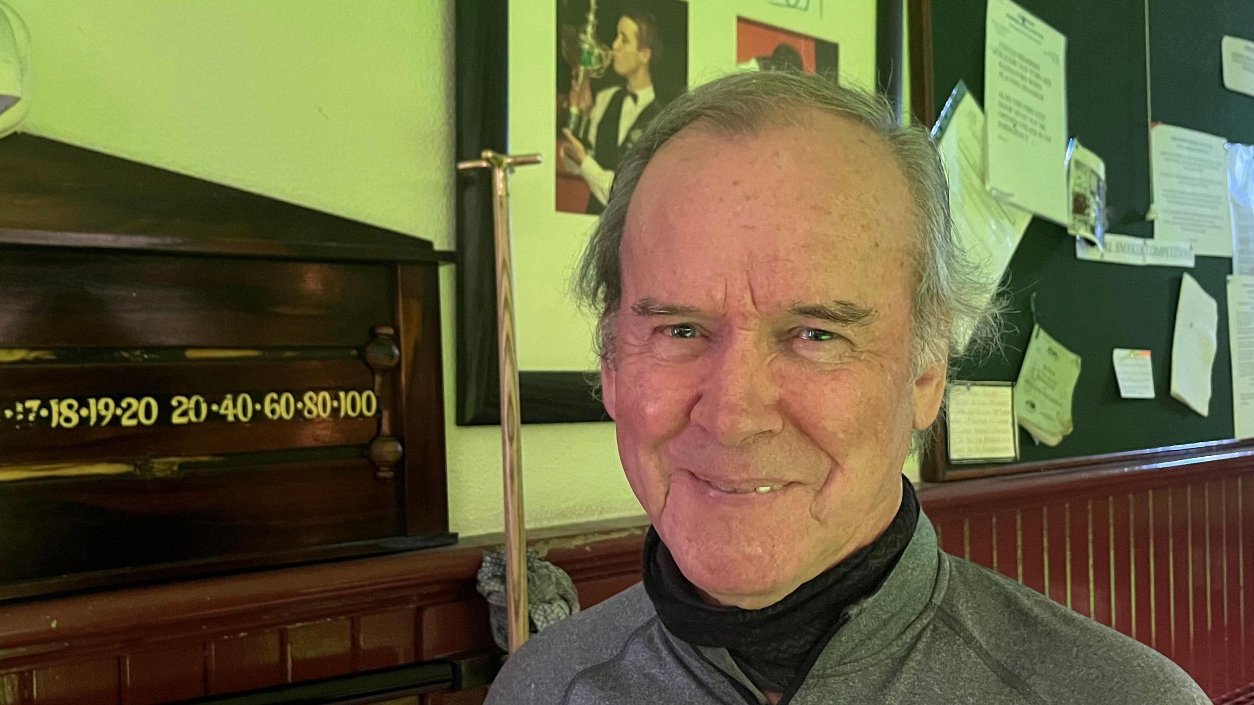 David Stephenson, wearing a grey top over a black polar neck. He is standing next to a snooker scoreboard. 