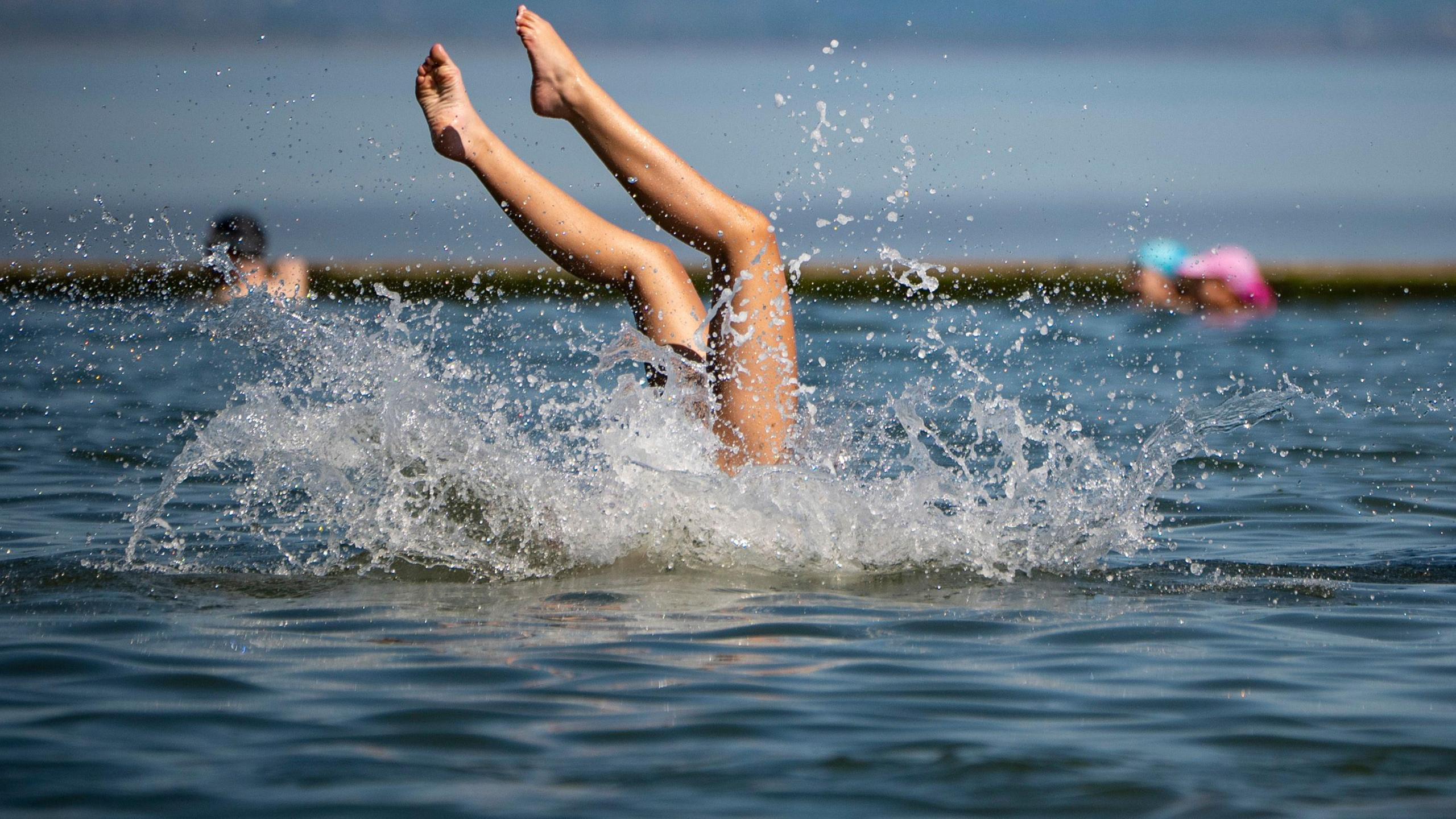 A woman's legs are seen sticking up out of the water from Clevedon Marine Lake as she dives in, creating a large splash, on a sunny summer day