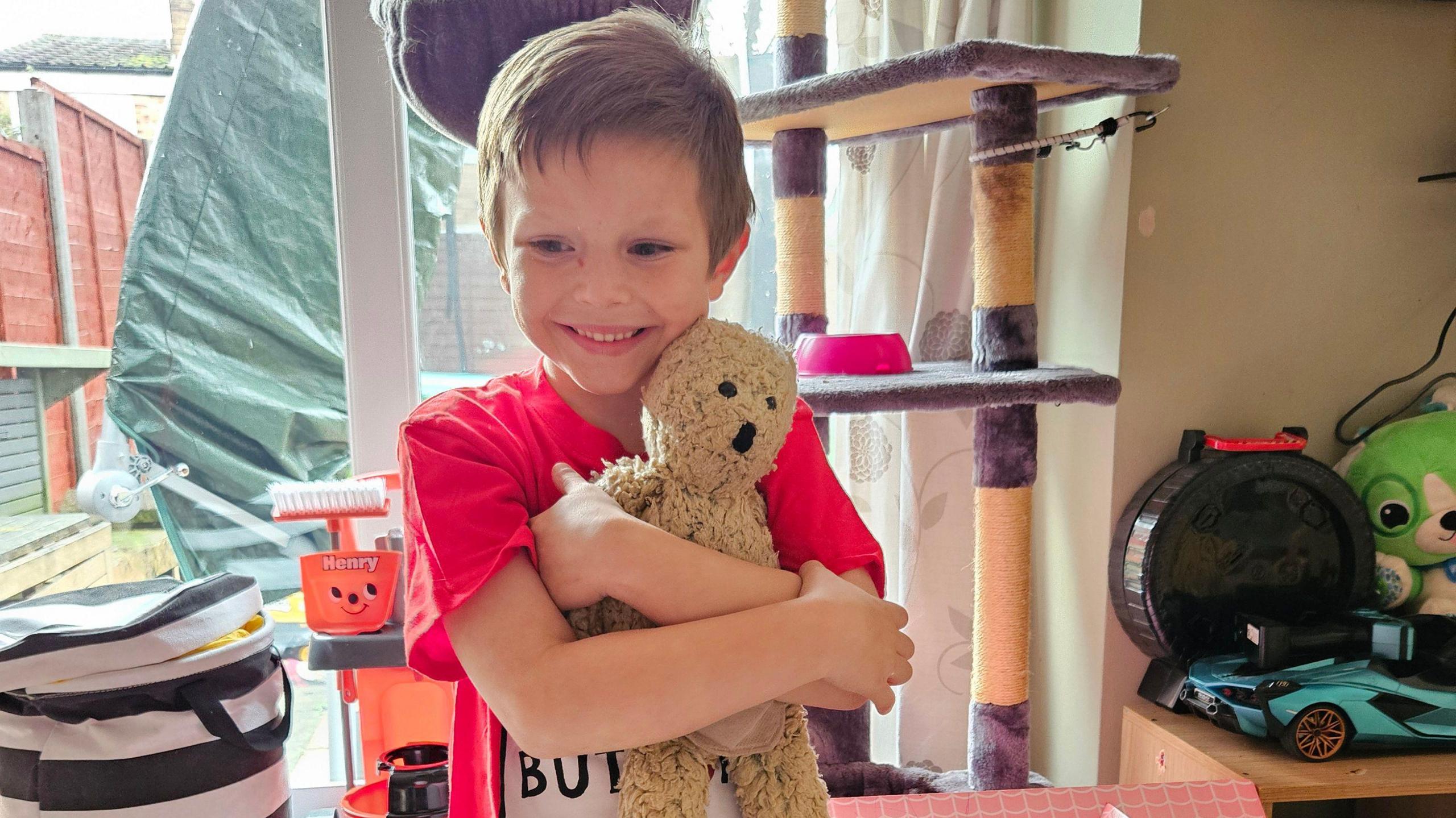 Riley smiling while cuddling his Buttony bear. He is wearing a red T shirt and has short brown hair.