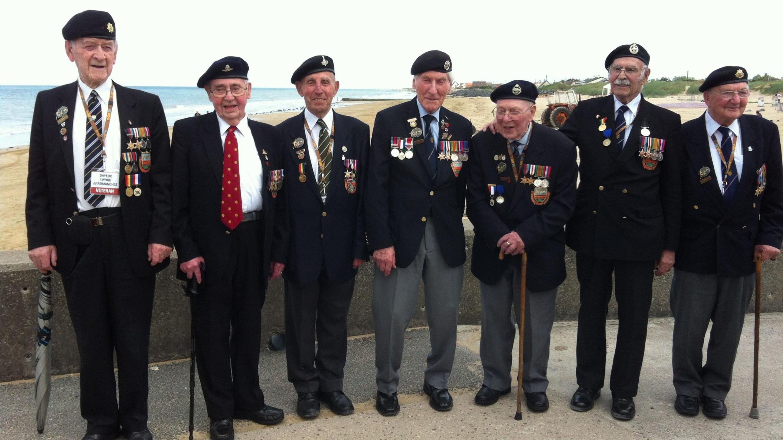 Seven older men wearing suits with many military medals and badges on them are lined up with a beach in the background. Some of them are holding walking sticks.