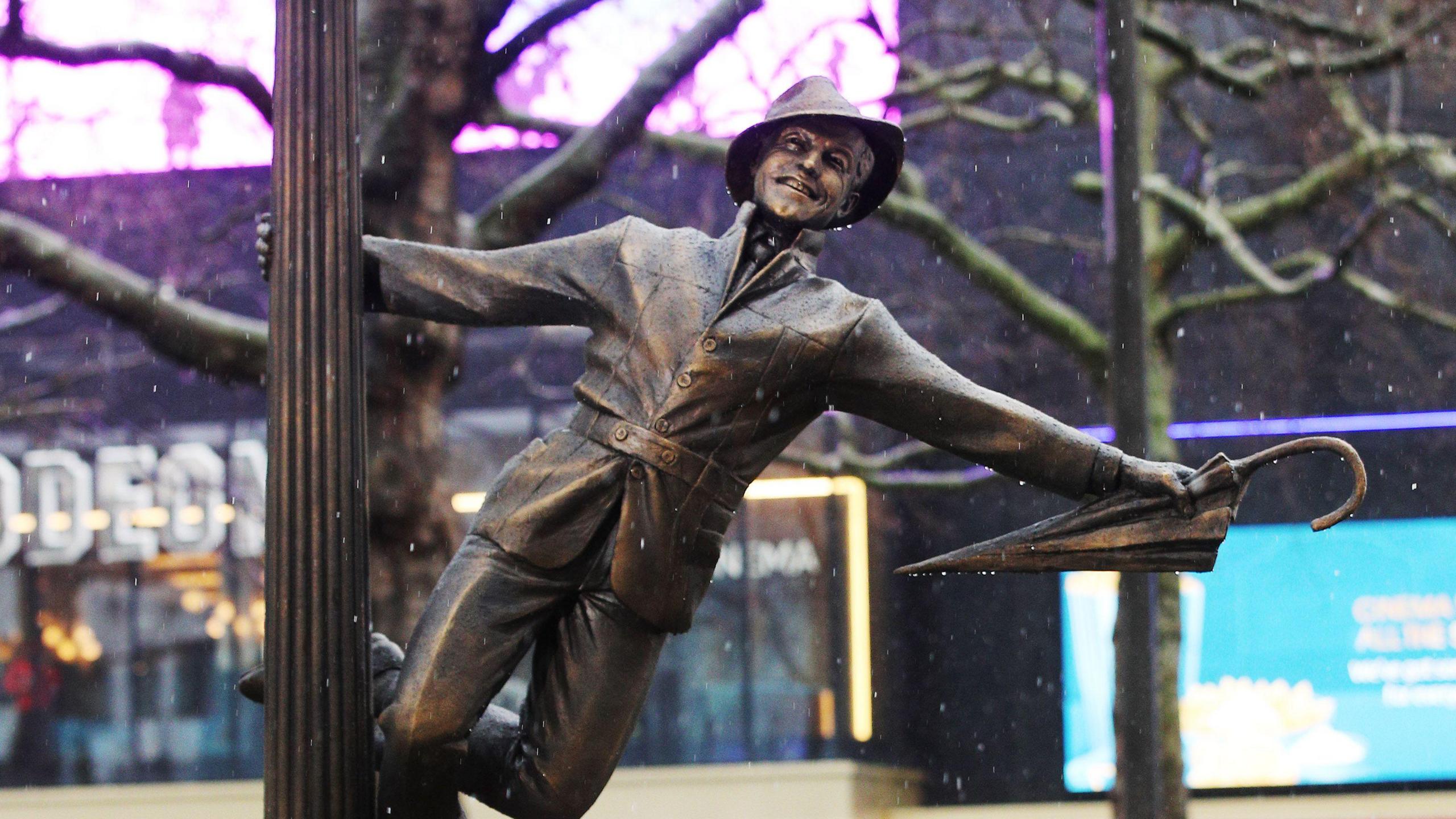 A statue of Gene Kelly hanging off a lamp post with an umbrella in hand in the classic pose from his film Singing in the Rain. The photo was taken when it was raining. A sign for the Odeon Leicester Square is in the background. 