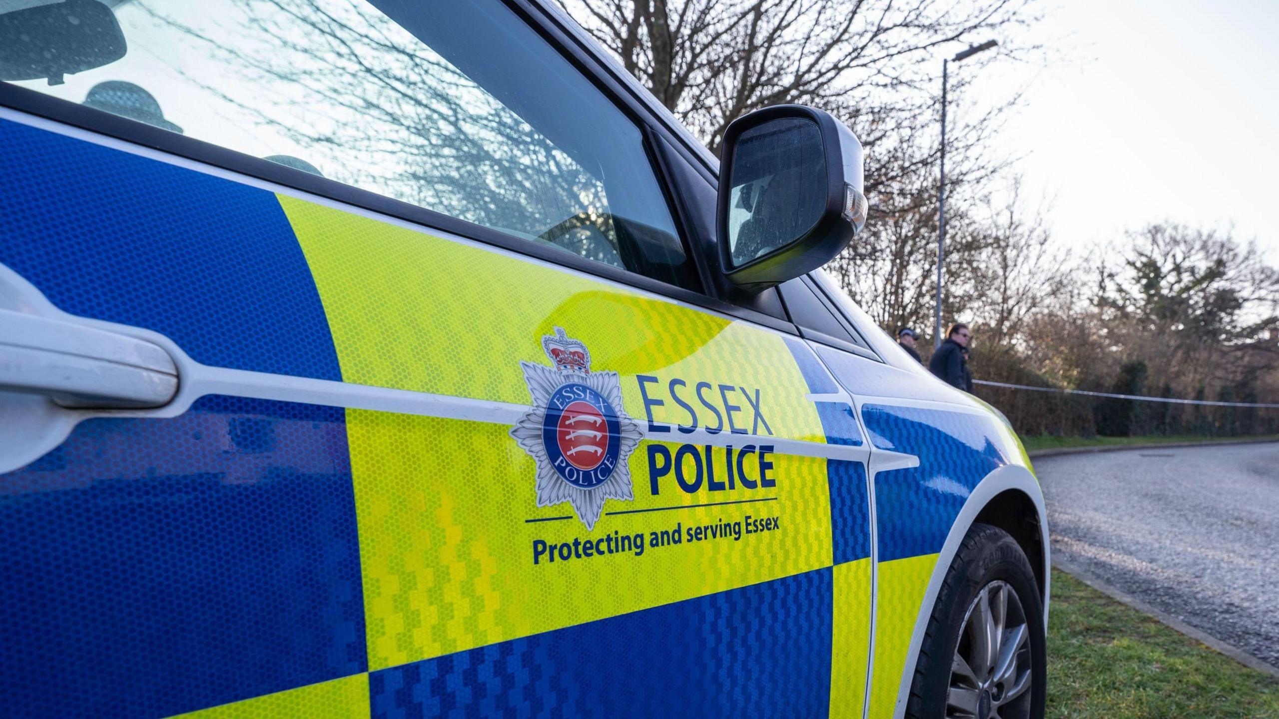 An Essex Police force car is parked on a grass verge next to a road. The road has a tape running across it to cordon it off to other drivers and pedestrians.  Two officers stand near the tape.