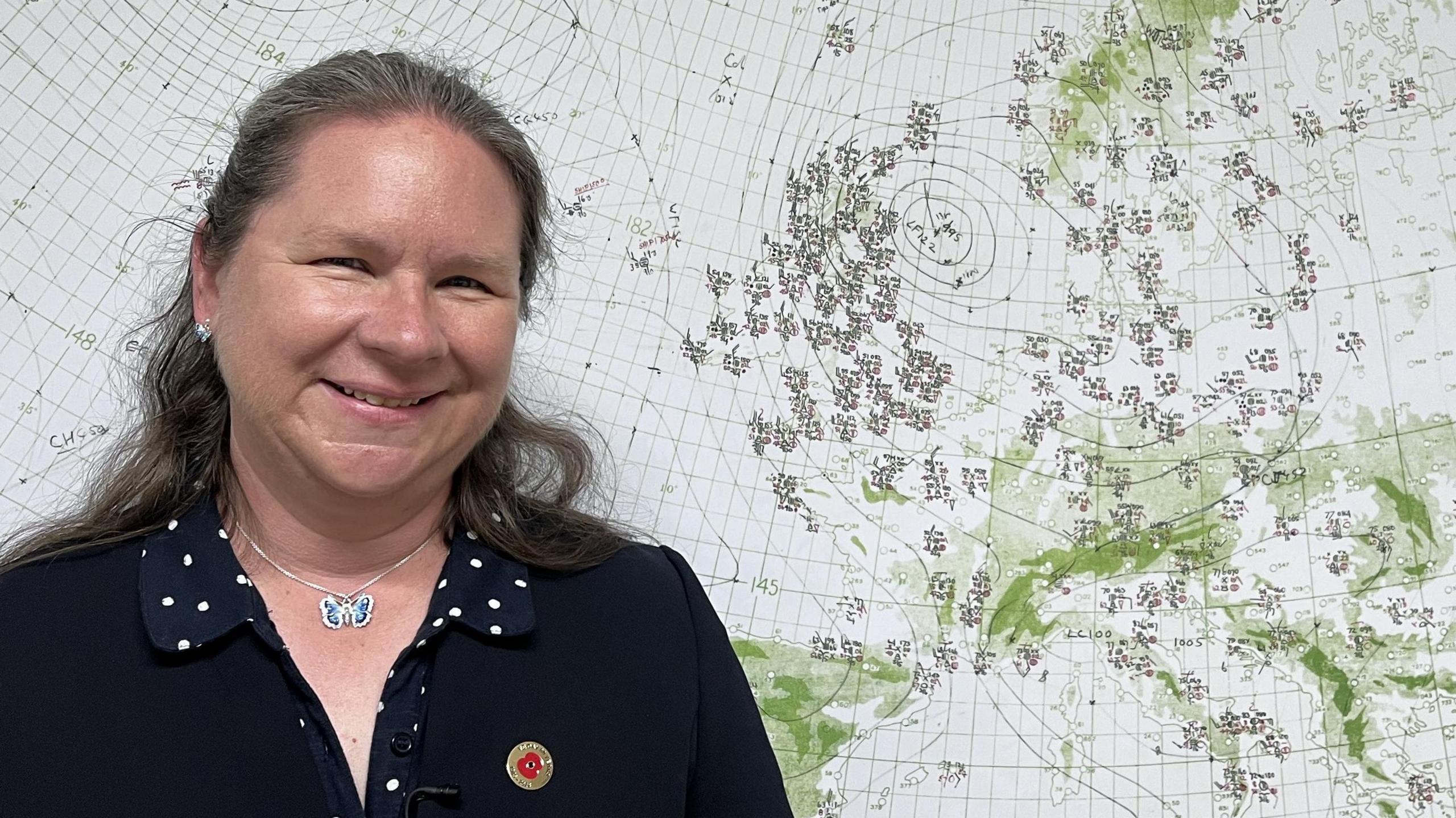 Dr Catherine Ross has dark grey long hair, and is wearing a dark jacket and a navy and white polka dot dress. She is smiling while standing in front of a hand-drawn weather chart from D-Day in 1944.