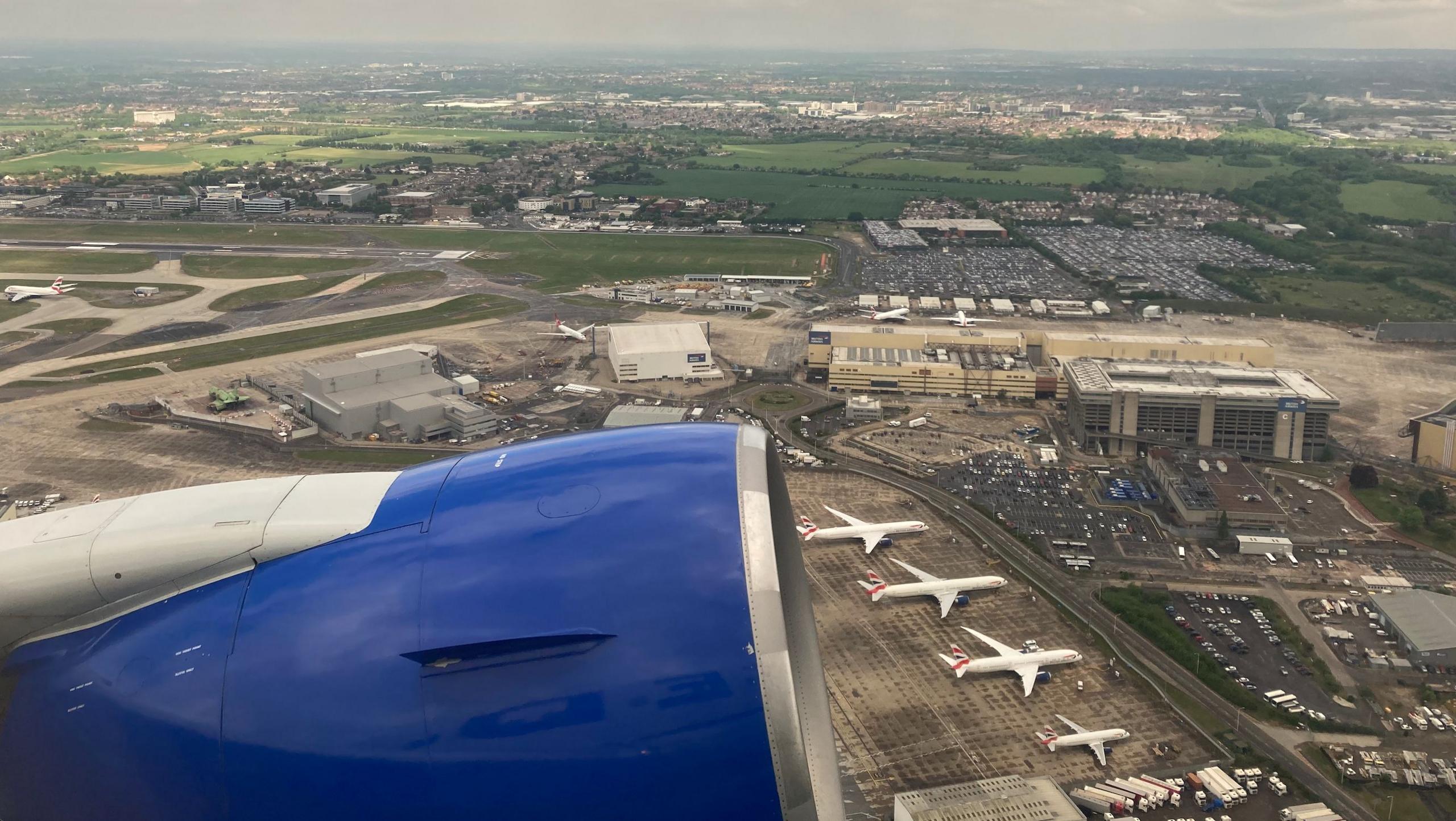 View from a plane shows blue engine of plane and four aircraft on tarmac outside low-rise terminal buildings.