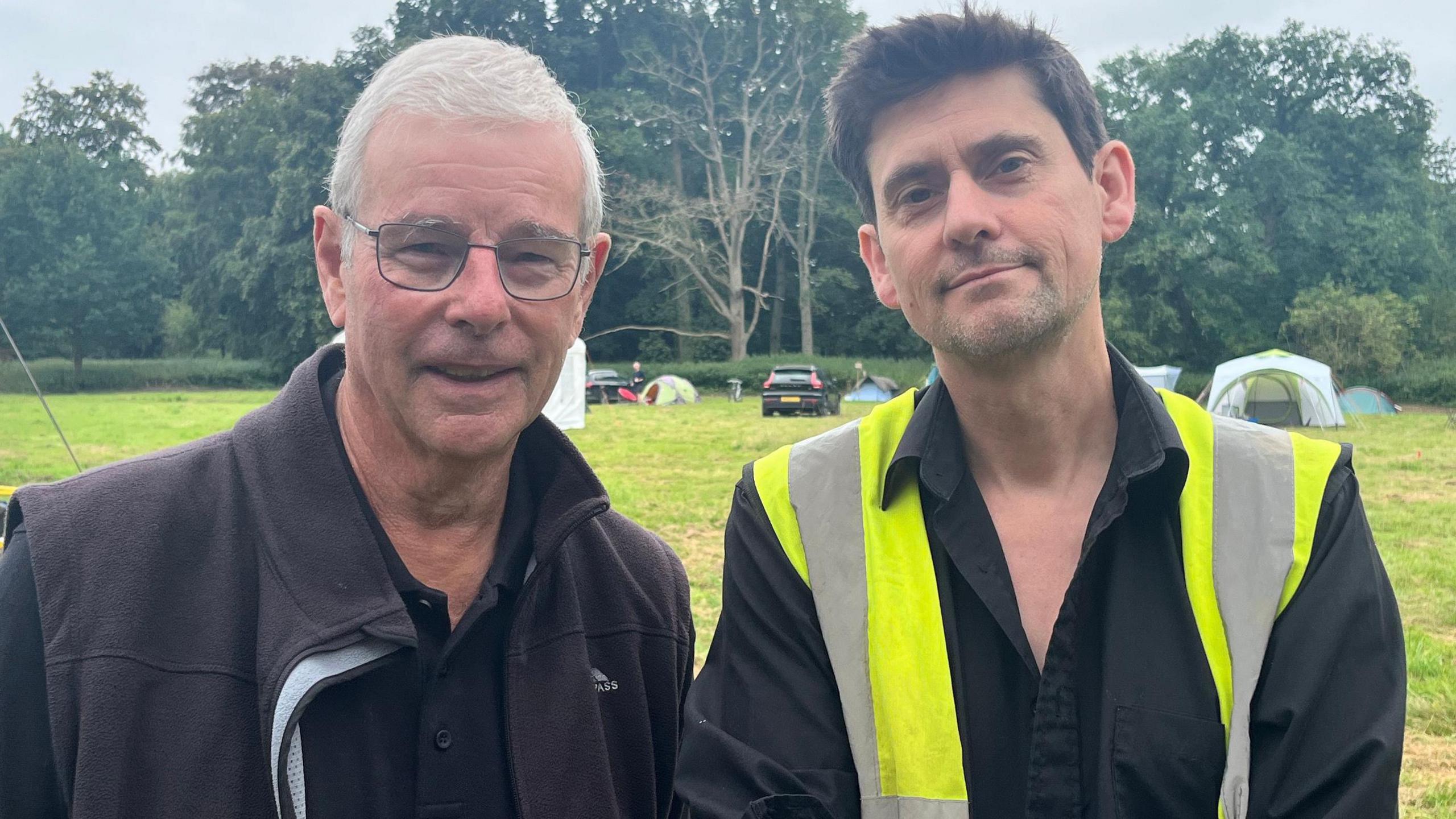 Mike Pinner is smiling at the camera and wearing a pair of glasses, dark polo shirt and fleece jacket. He is standing next to Giles Emery, who is wearing a high vis jacket and black shirt, which is unbuttoned at the neck. They are standing in a field, with tents, cars and trees in the background.