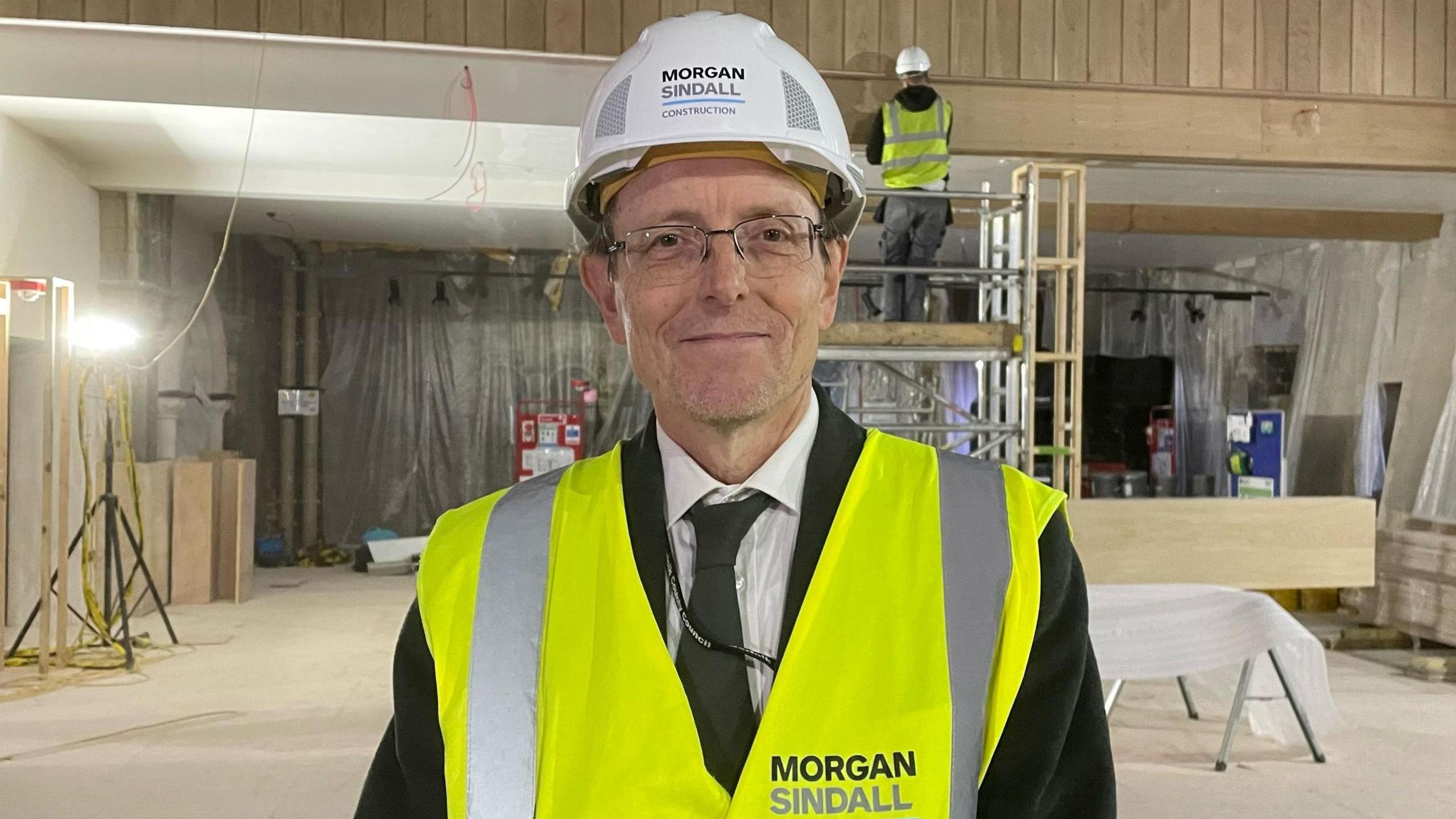 Dr Robin Hanley is dressed in a suit with a high-vis jacket on top. He has a white helmet and glasses. He is standing in Norwich Castle. A workman is stood on scaffolding behind him.