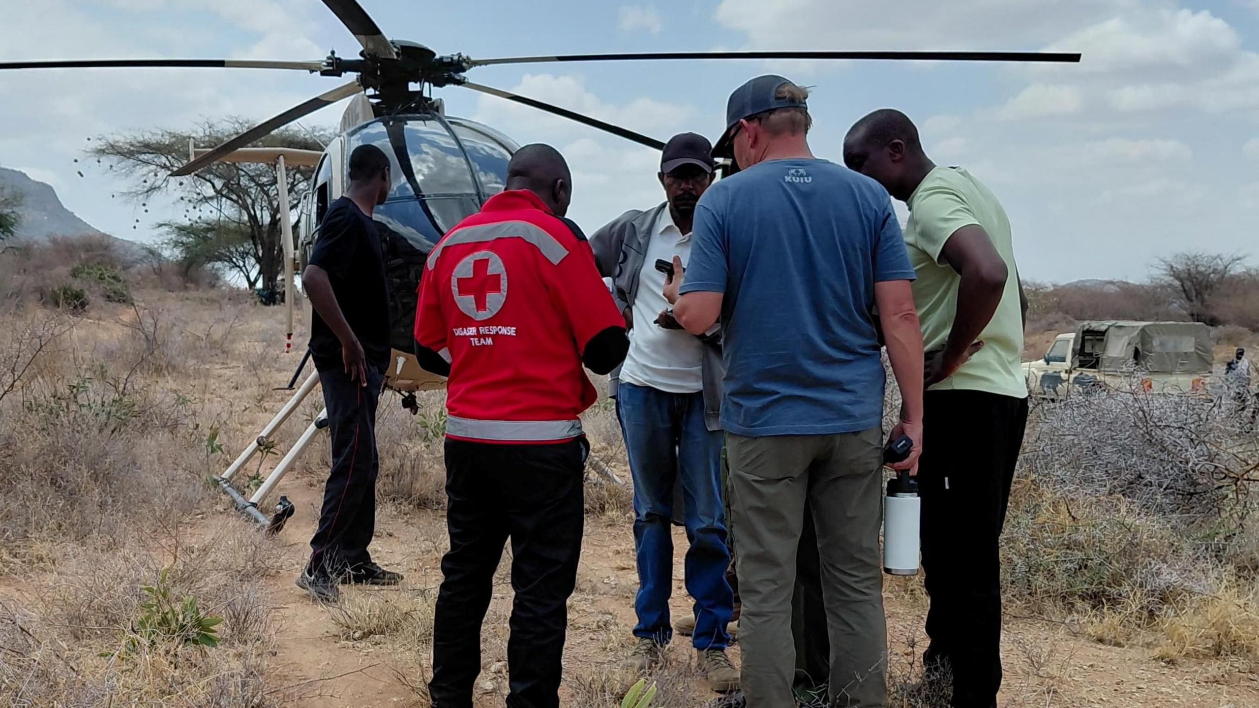 Five men stand on dusty arid dirt with a helicopter in the background. One of them has a Red Cross top on.