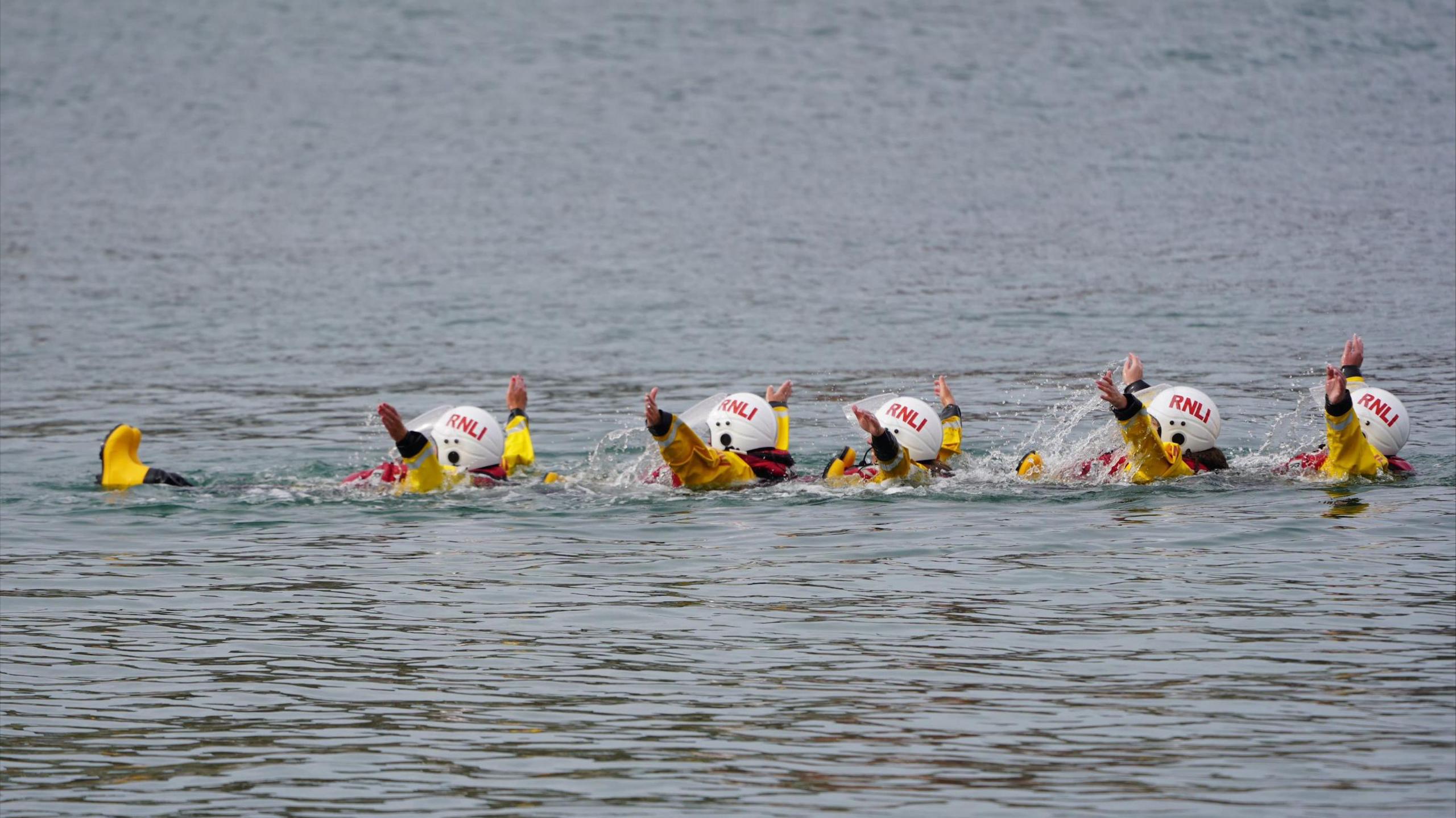 RNLI Cullercoats crew wear yellow and black uniform and red lifejackets. They are doing a team exercise in the sea and are floating with their hands up in the air.