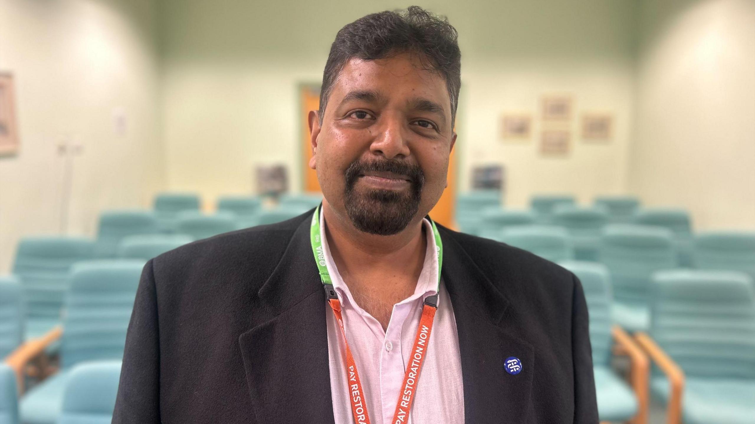 Prakash Thiagarajan smiling. He is wearing a dark suit jacket and pink shirt and standing in front of some teal green chairs.