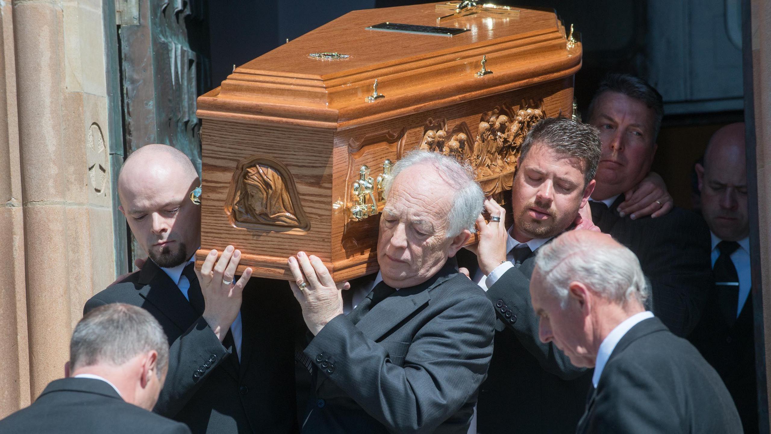 A group of men in black suits are carrying a wooden coffin 