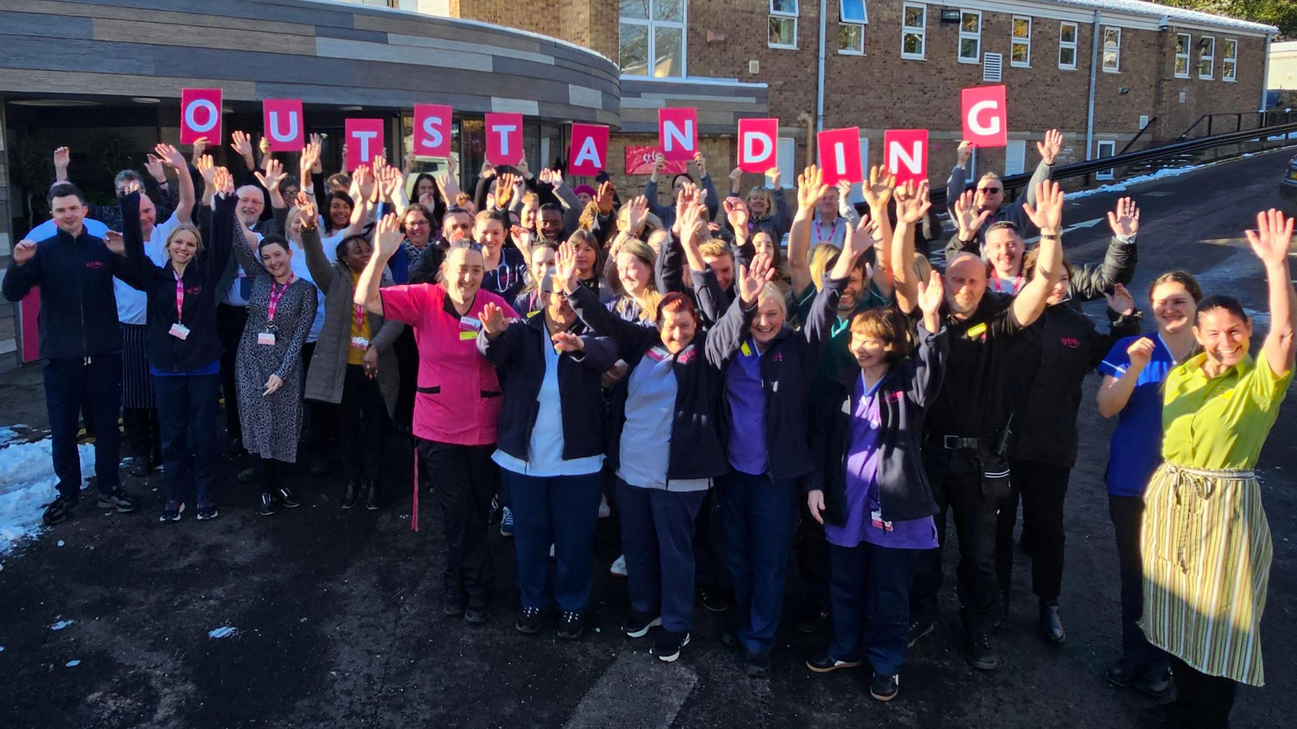 A group of approximately 30 people lined up in three rows facing the camera, which their arms in the air smiling.  Some hold up individual letter posters which combine to make the word outstanding