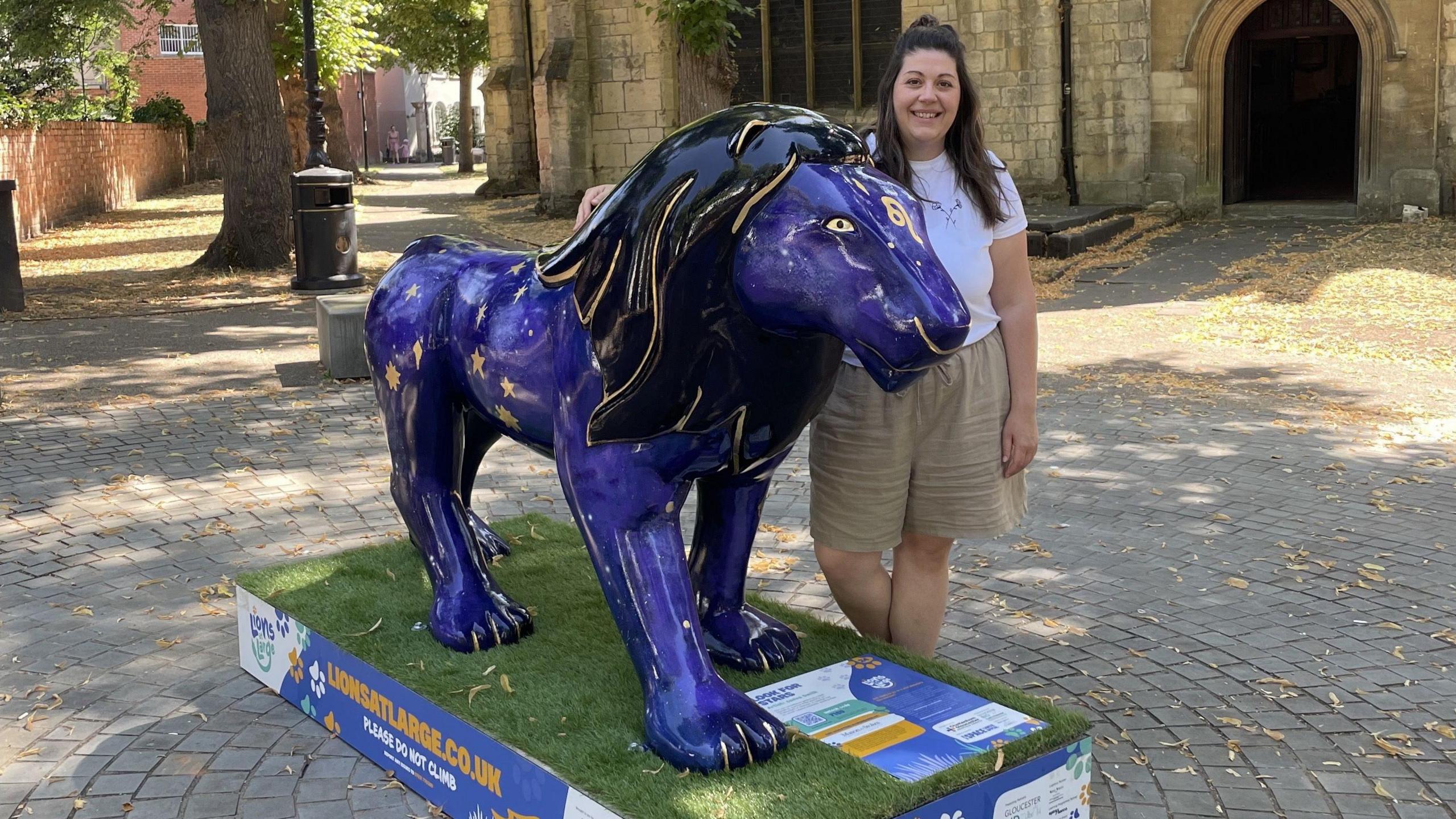Artist Janine Smith is standing next to a purple lion with a black main and gold streaks running through it. Janine is wearing a white t-shirt with brown shorts standing in front of a church with her hand on it.
Location: Minster Exchange