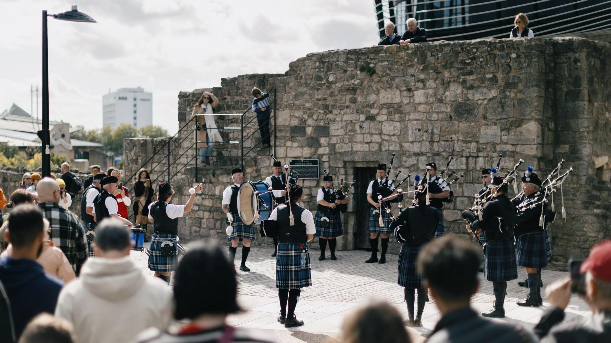 A band of bagpipers play next to a medieval wall.