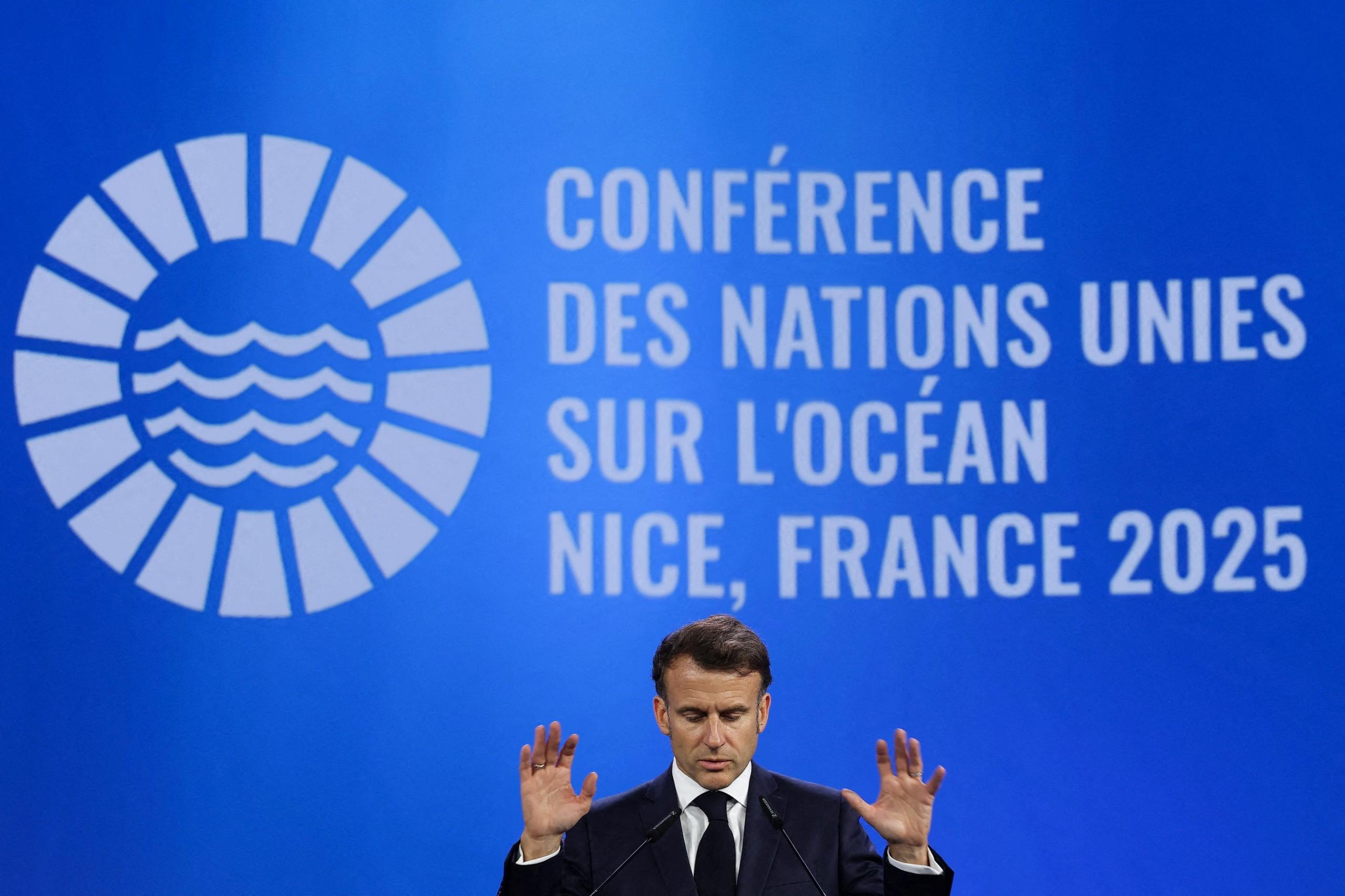 President Macron is standing in front of a lecturn in a black suit, he is looking down with his hands raised and palms facing forward. There is a blue backdrop that reads "UN Ocean Conference, Nice France 2025" in French