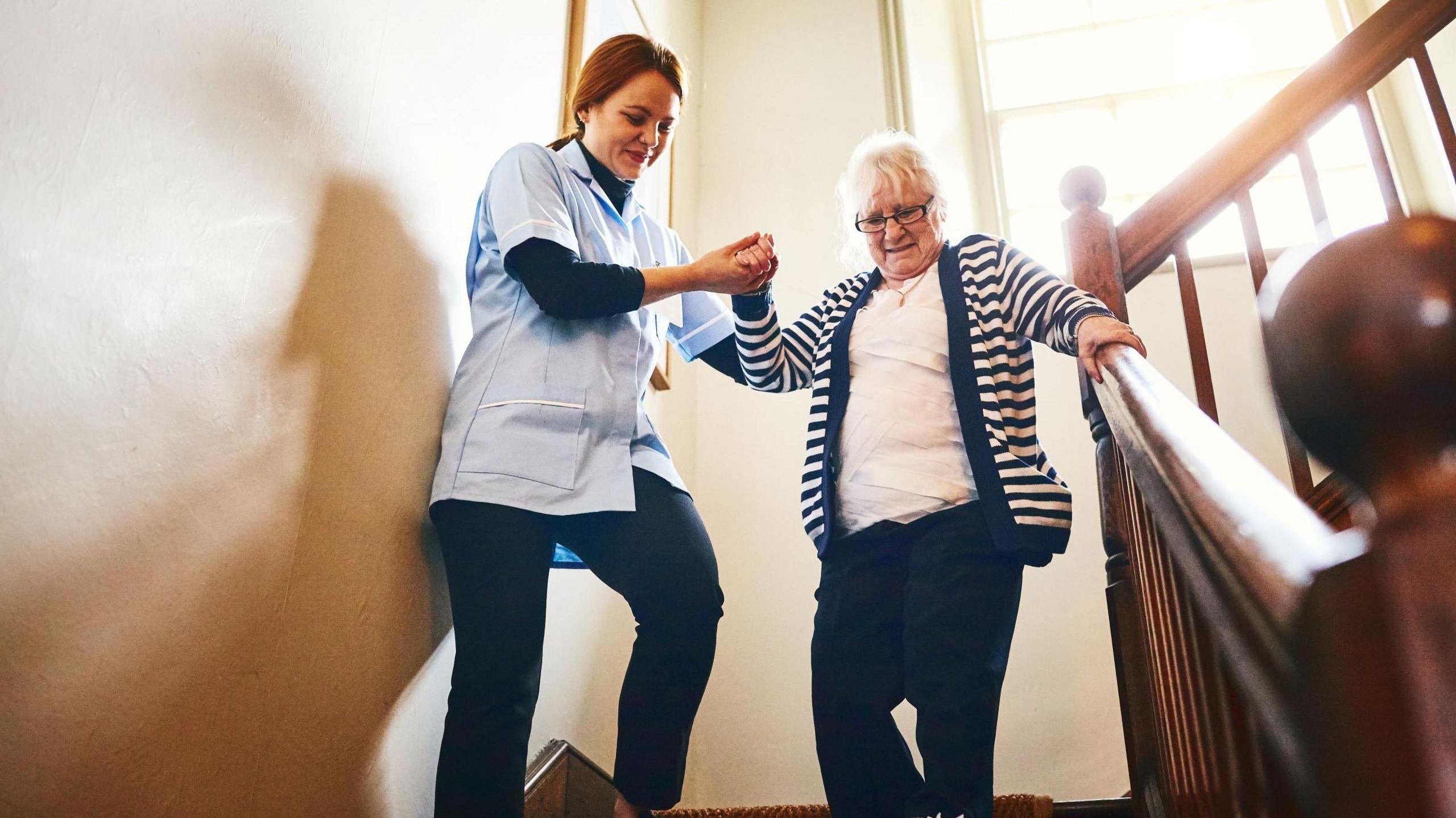 A smiling nurse helps an older woman walk down stairs