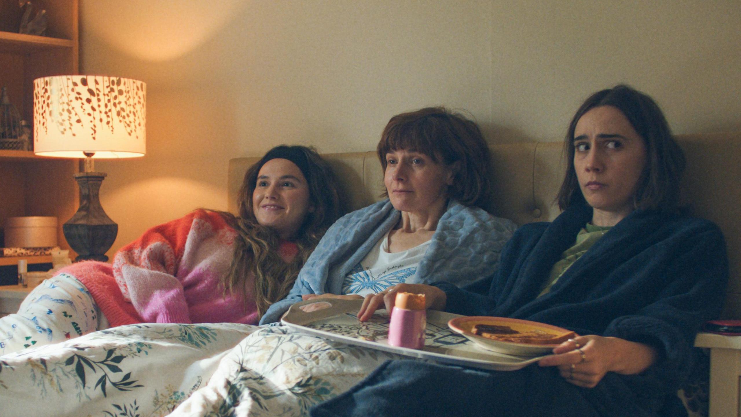 Left to right: In bed next to each other are Billie (Lizzie Davidson) wearing a pink jumper, Deb (Louise Brealey) wearing a white top and a blue dressing gown, and Josie (Kat Sadler) wearing a green top and a blue dressing gown and holding a tray with toast and a boiled egg on.